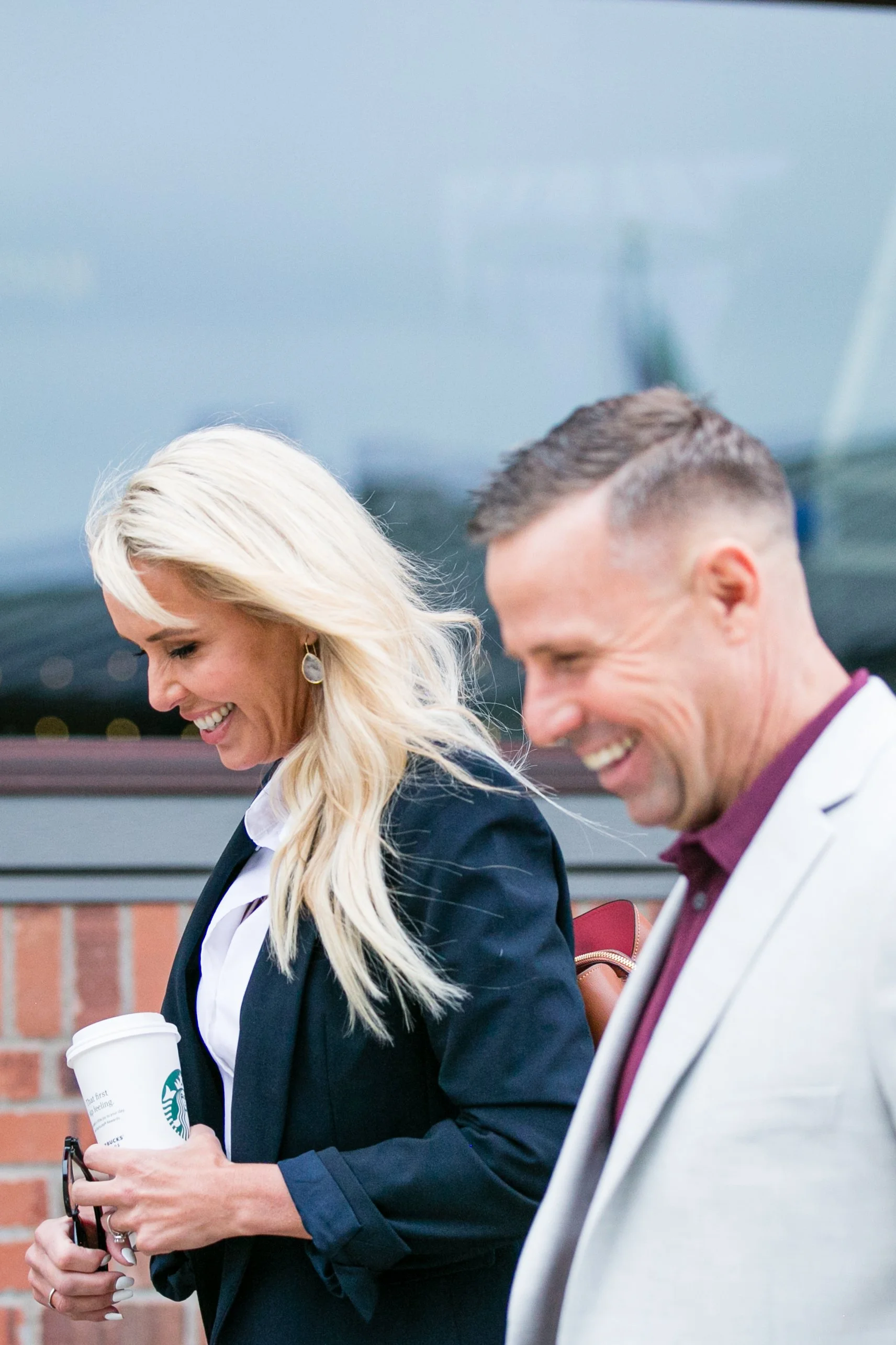 Two smiling people, a woman with long blonde hair holding a Starbucks coffee cup and a man with short hair wearing a white blazer, walking outdoors.