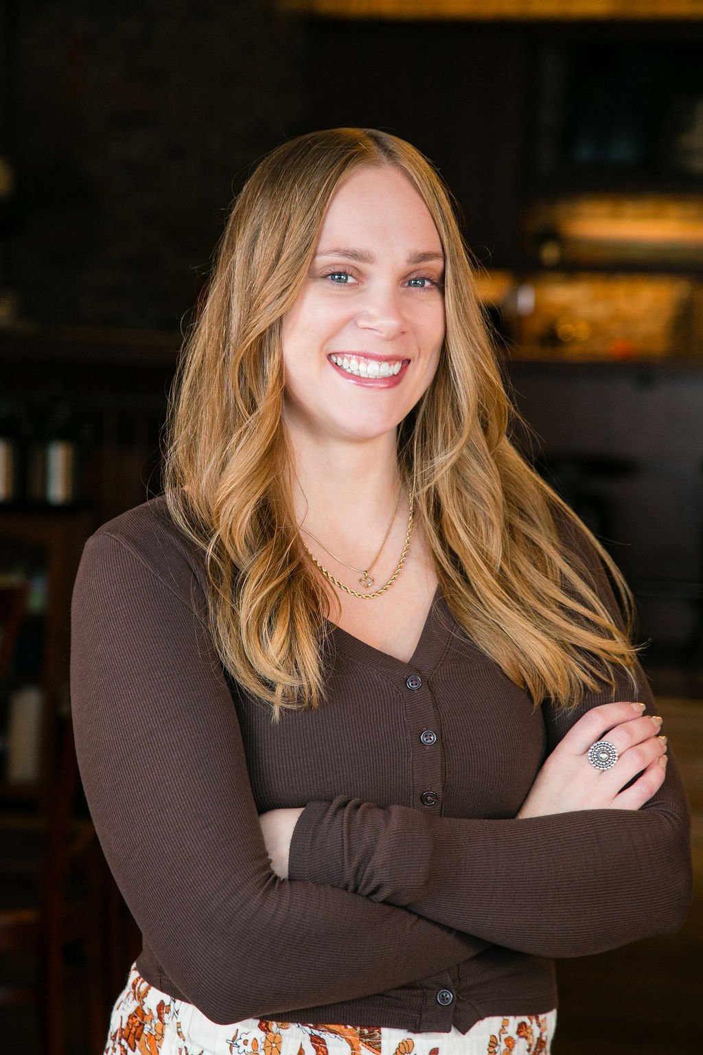 A young woman with long blonde hair smiling, wearing a brown cardigan, layered necklaces, and a ring, standing with arms crossed indoors.