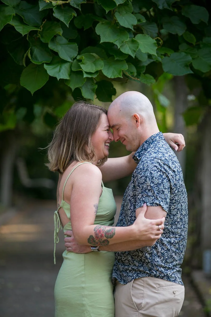 A man and woman are smiling and touching foreheads, standing close together outdoors with green foliage in the background.