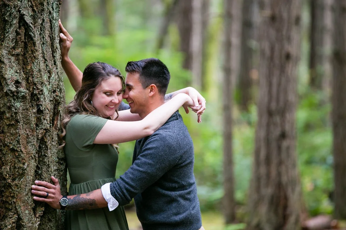 A couple embracing and smiling with foreheads touching in a wooded forest, with trees in the background.