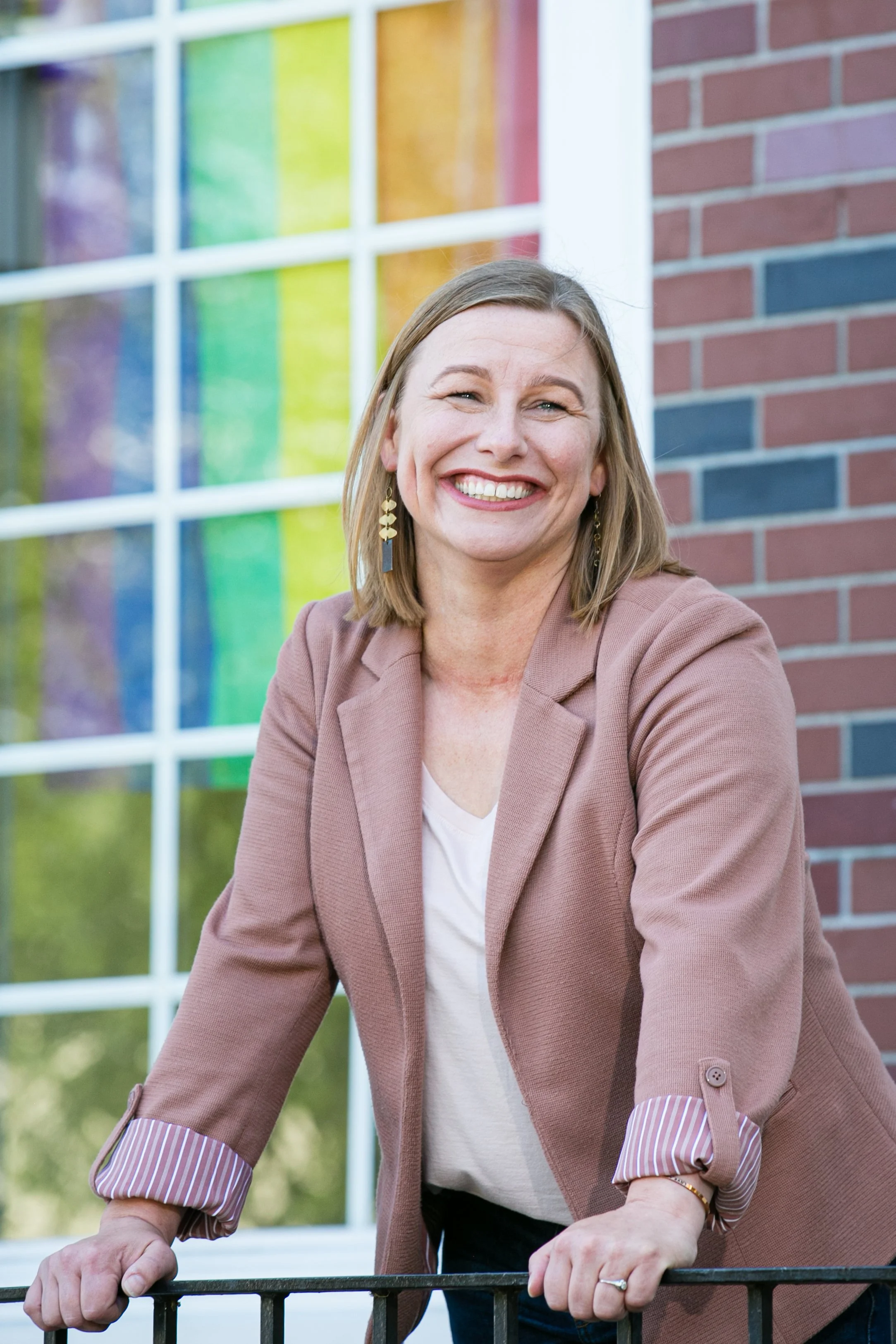 A woman with shoulder-length blonde hair smiling, wearing a light brown blazer with striped cuffs and earrings, leaning on a black railing in front of a building with a colorful mosaic window and brick wall.