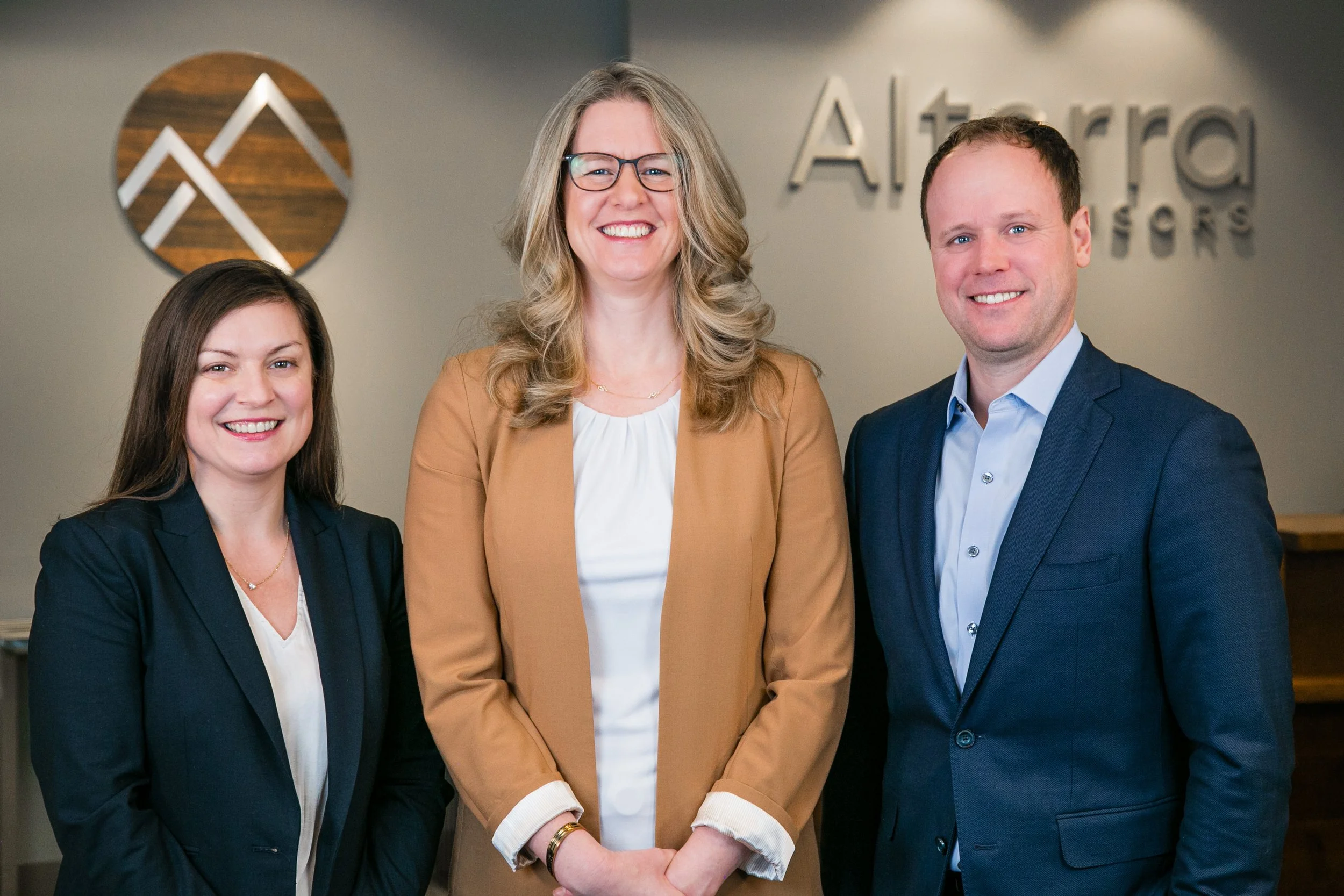 Three professionals standing together in front of a wall with the logo 'Altierra Attorneys'. The woman on the left has long brown hair and is wearing a black blazer and white blouse. The woman in the middle has long blonde hair, glasses, and is dress