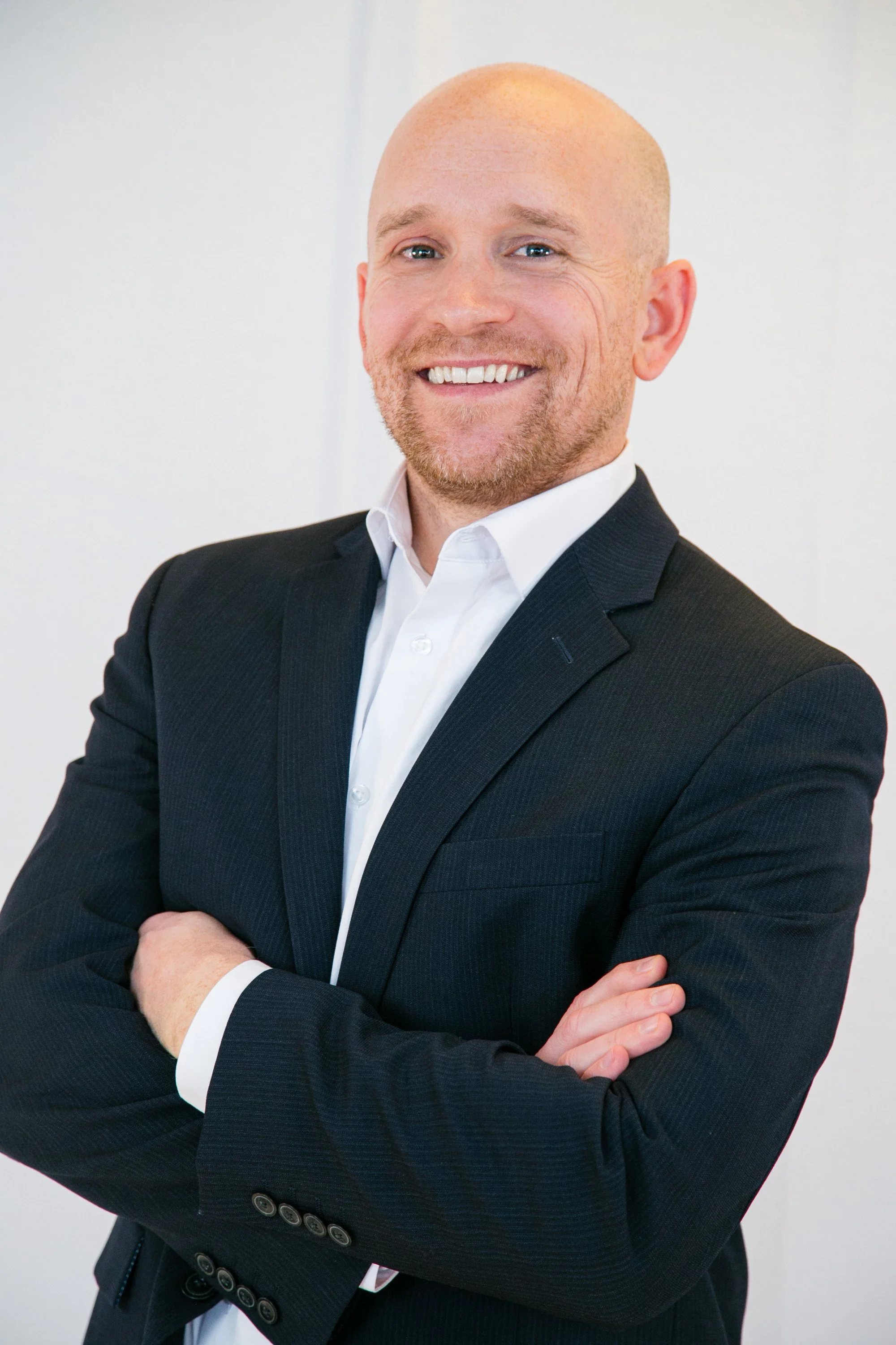A man smiling with arms crossed, wearing a black suit and white shirt in a professional setting.
