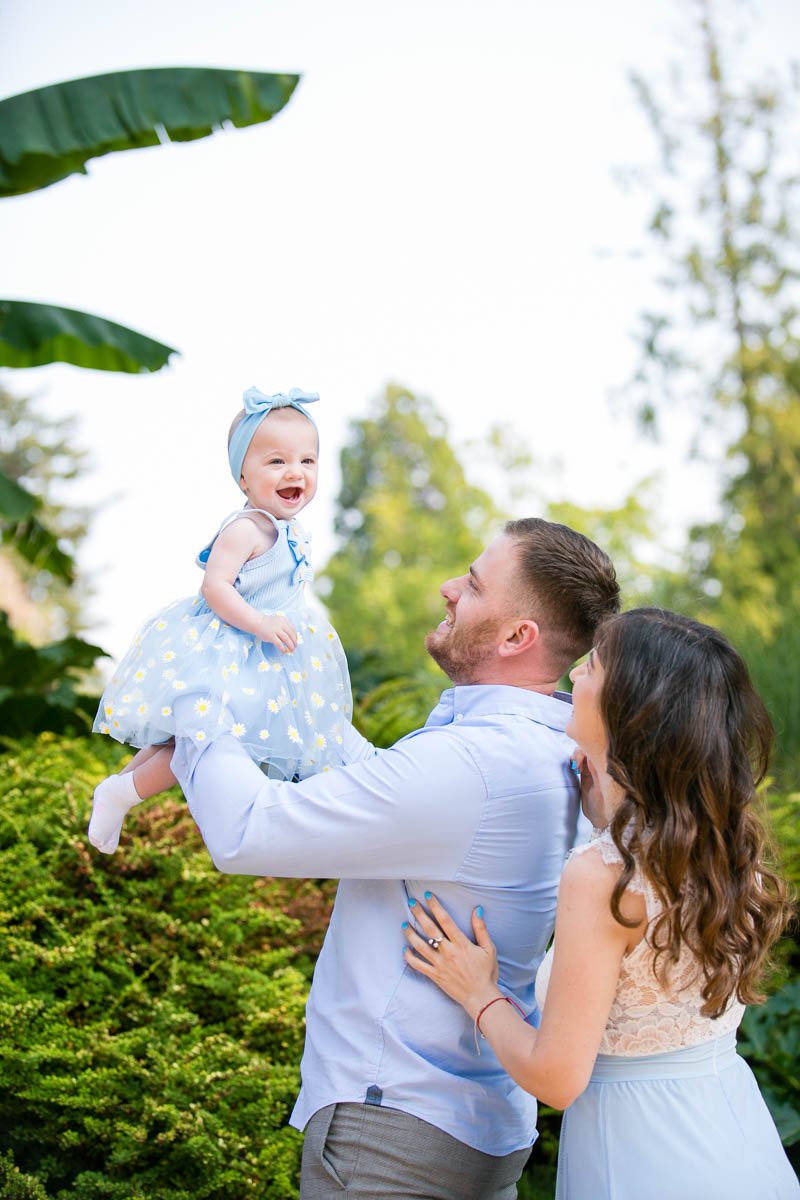 A man lifting a smiling baby girl with a headband and dress, while a woman stands nearby outdoors in a garden setting.