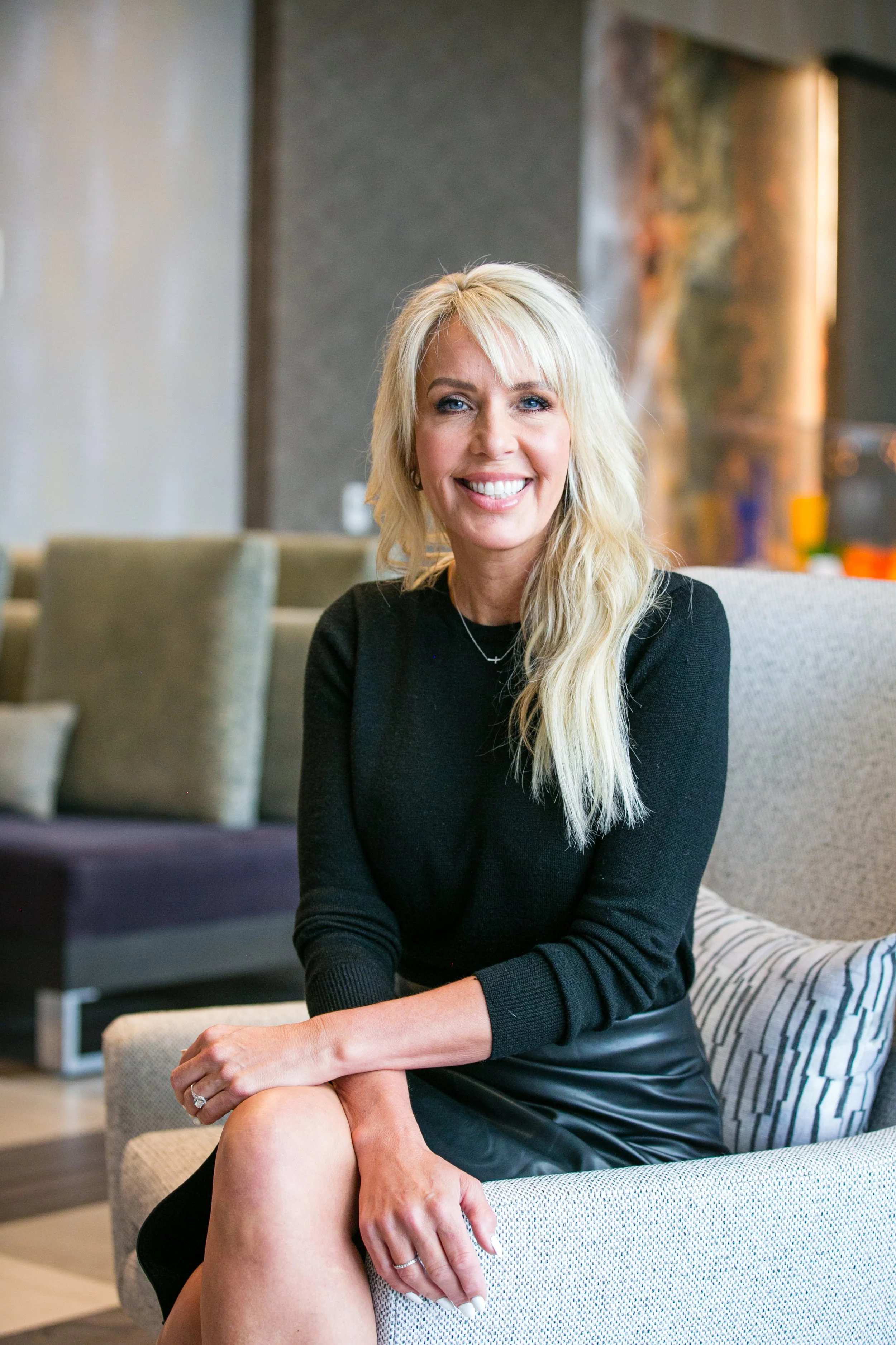 A woman with long blonde hair smiling, sitting on a light-colored sofa in a modern indoor setting.