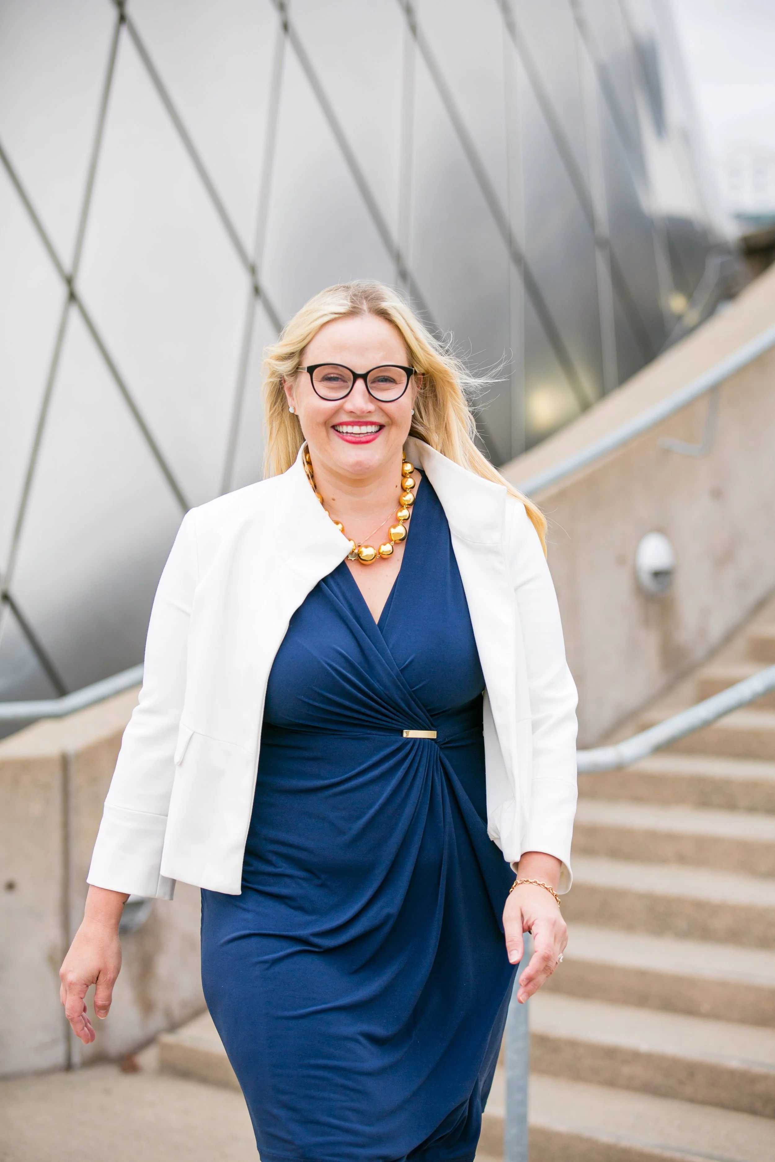 Smiling woman with blonde hair, glasses, blue dress, white blazer, and gold jewelry walking outdoors near stairs and modern building.