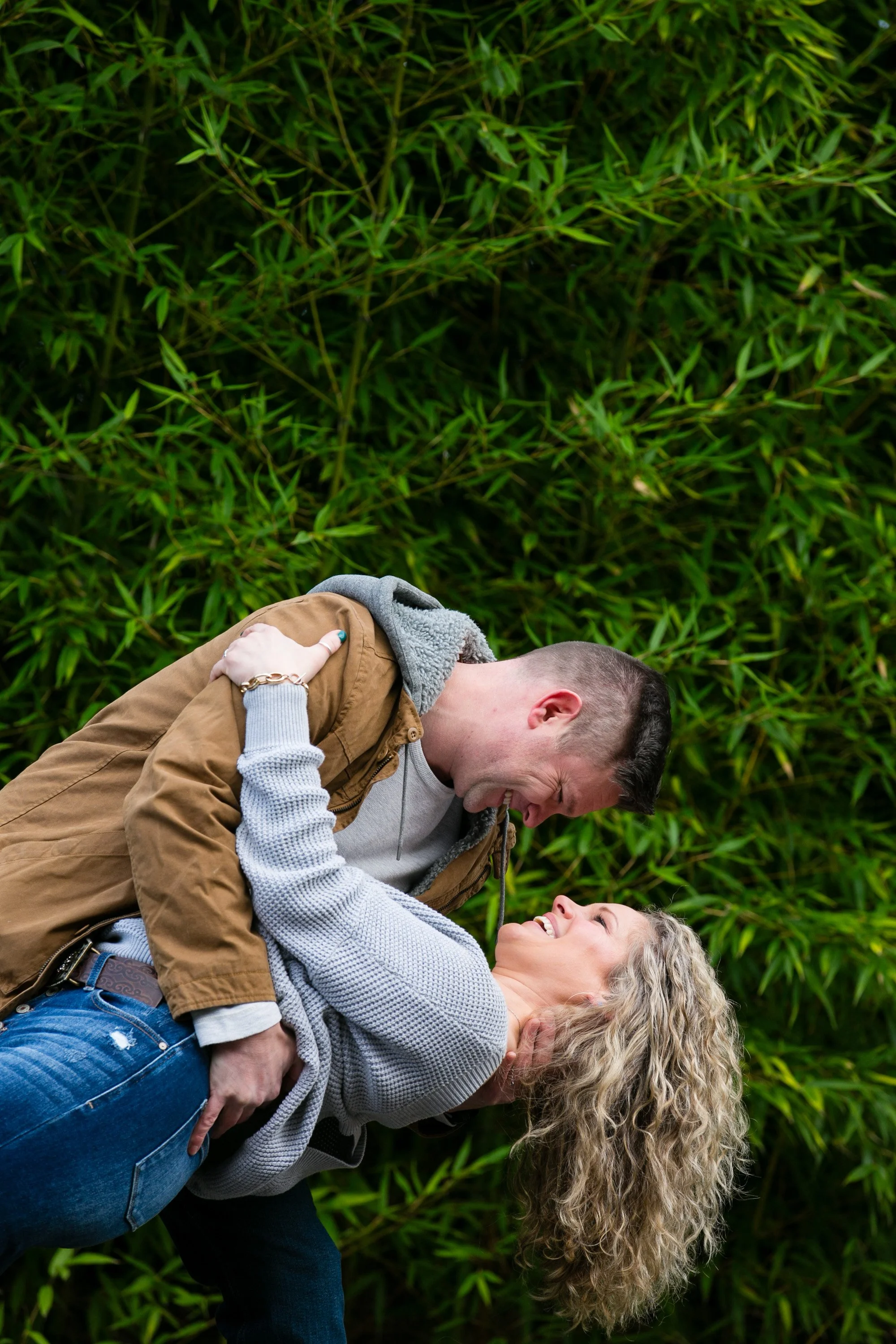 A young man is holding and smiling at a woman who is leaning back, surrounded by green foliage.