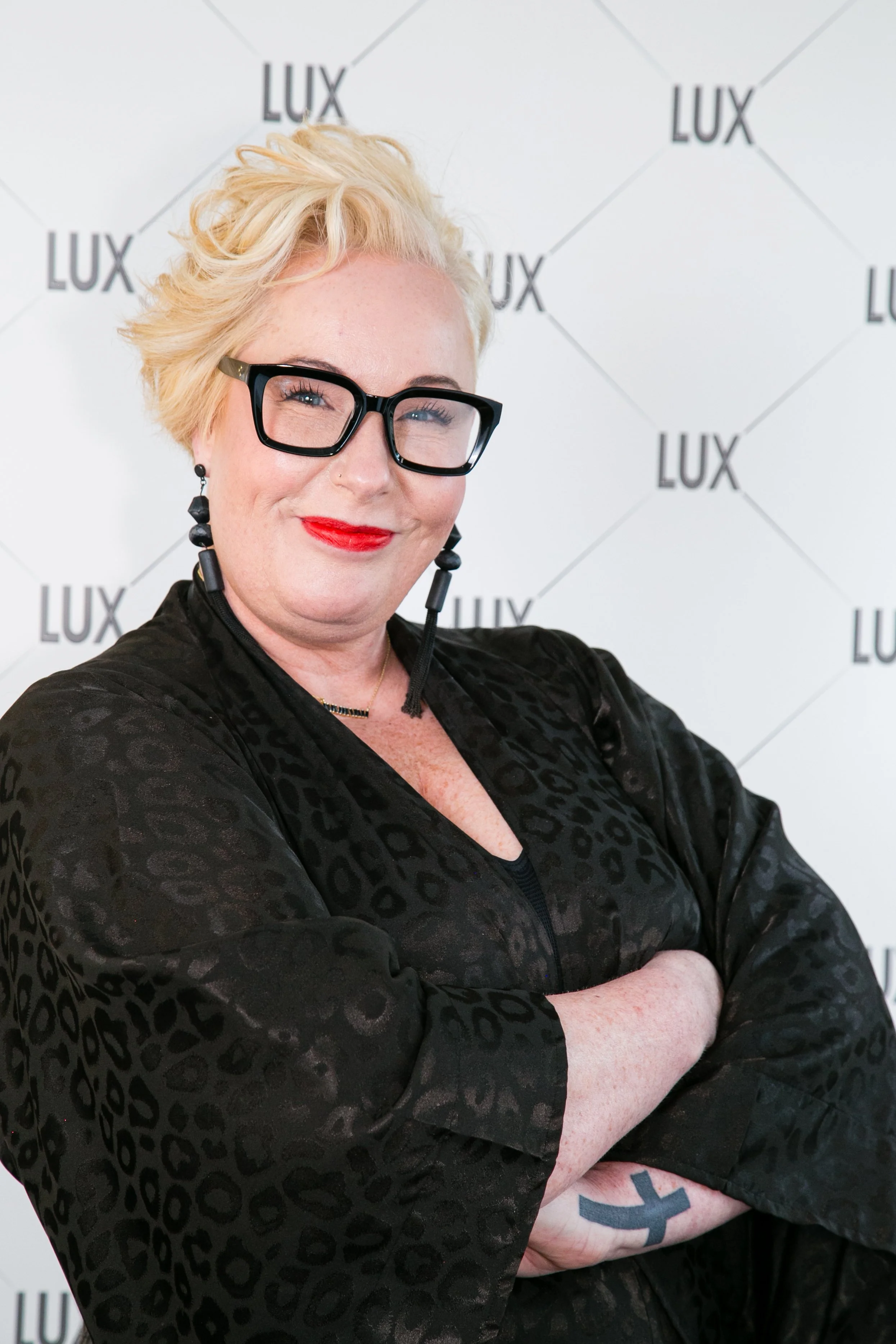 Woman with short blonde hair, black glasses, red lipstick, and black earrings, standing with arms crossed in front of a background with the word 'LUX' repeated multiple times.