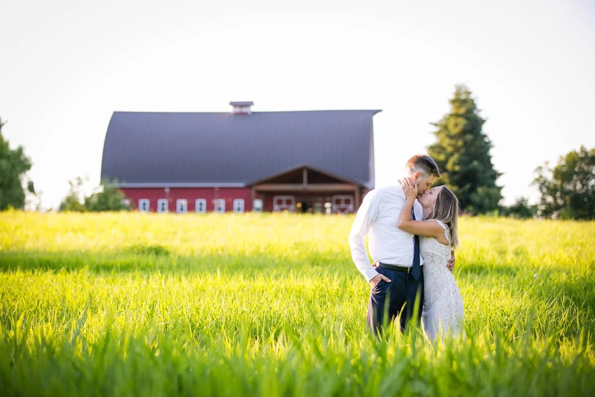 A couple kissing in a grassy field with a red barn in the background during sunset.
