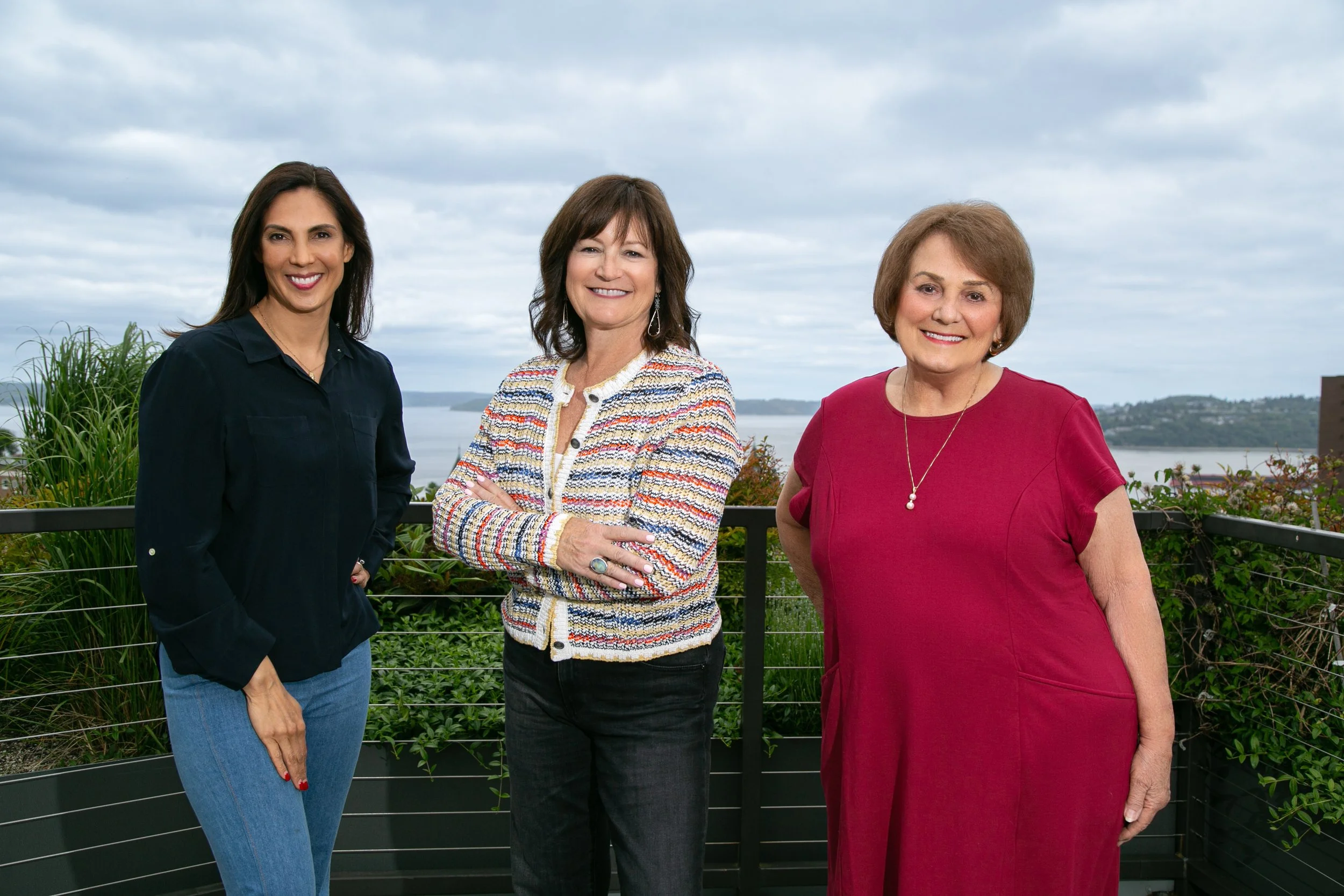 Three women standing outdoors on a balcony with a scenic view of water and hills in the background, smiling at the camera.