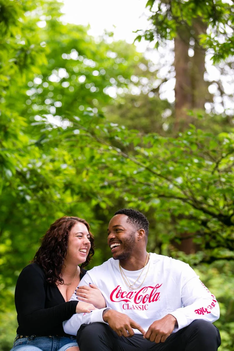 A man and woman sitting closely outdoors in a lush green environment, smiling and laughing together.