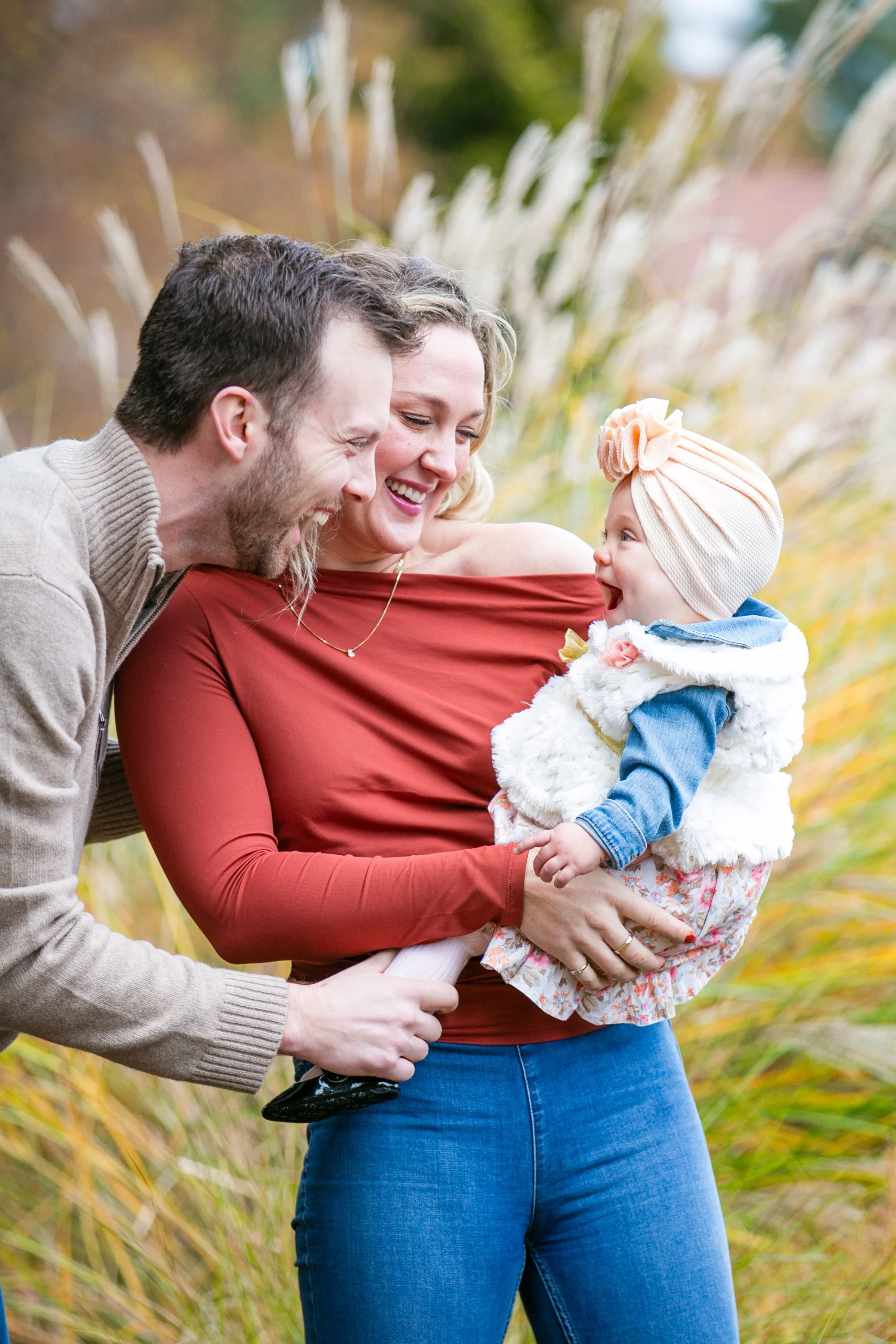 A family of three outdoors, smiling and laughing while a woman holds a baby girl.