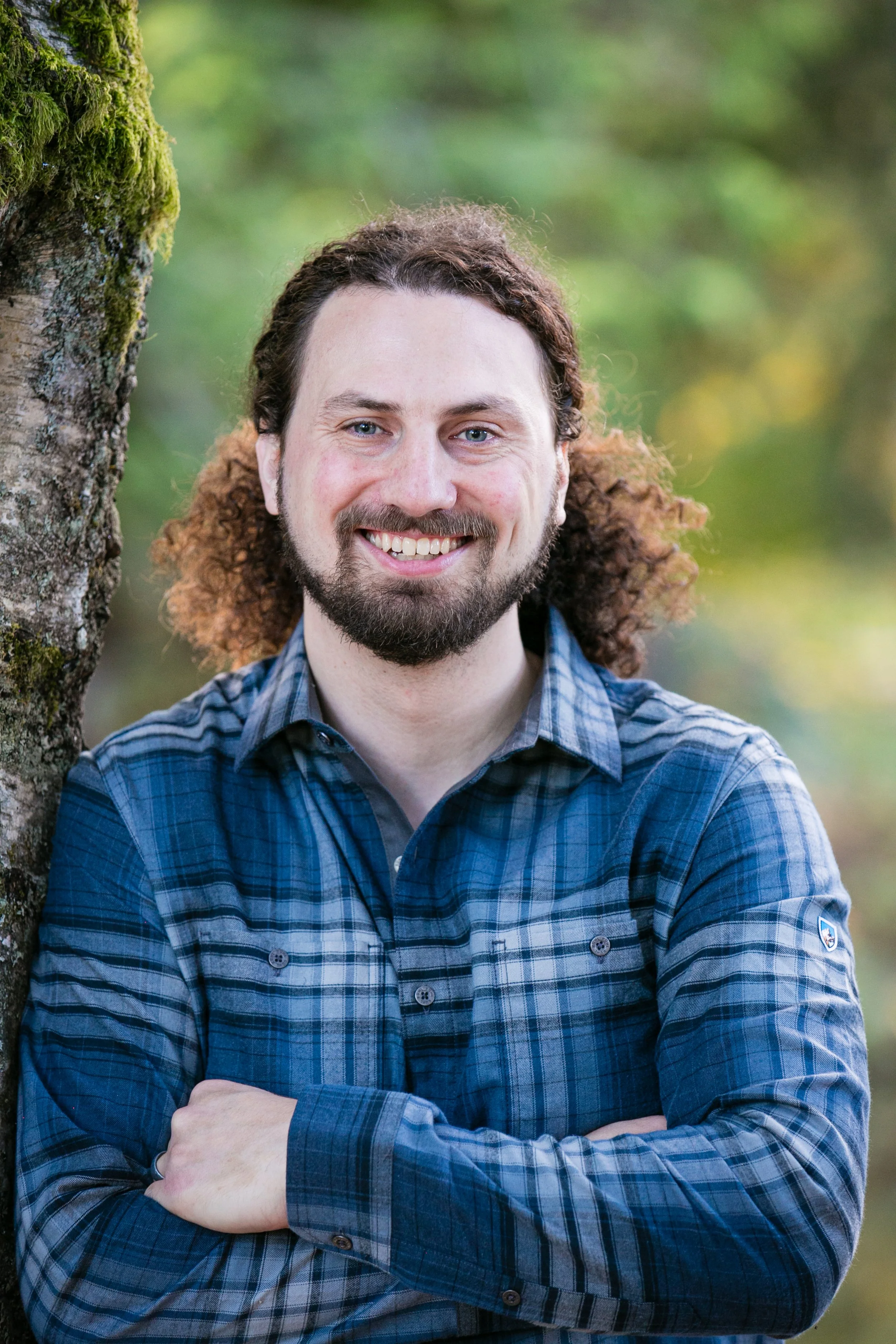 A man with curly hair and a beard smiling, standing outdoors with a tree trunk in the background.