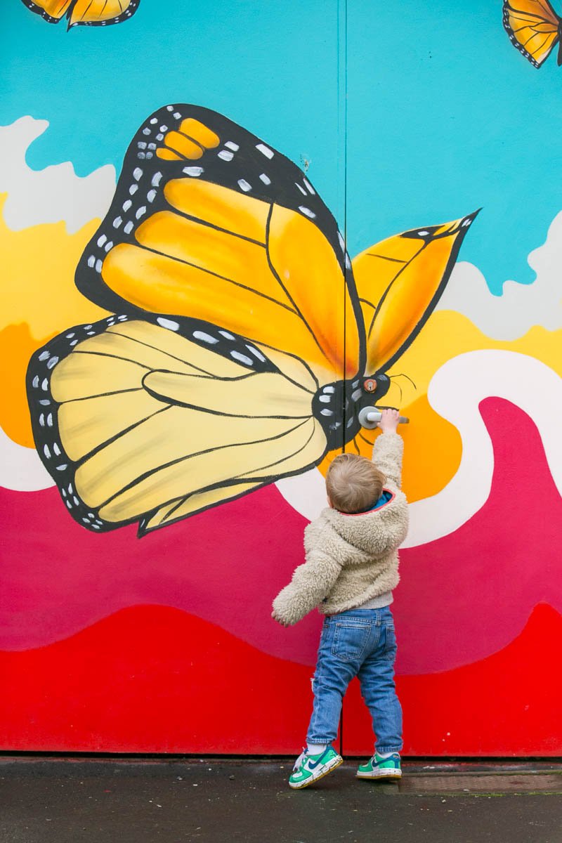 A young boy drawing on a colorful mural of a monarch butterfly with a white marker. The mural features bright backgrounds with clouds and landscape, emphasizing the butterfly's vibrant colors.