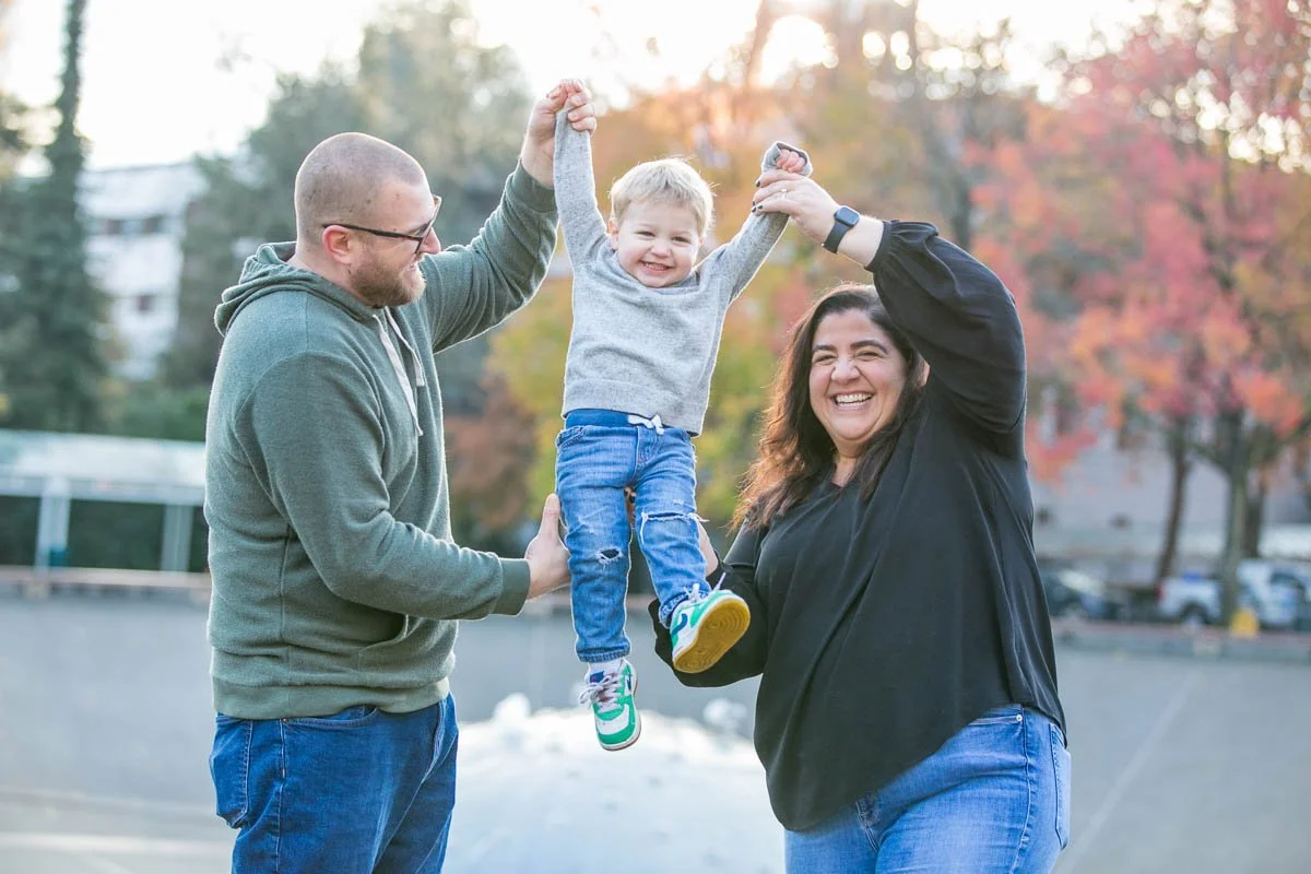 A happy family of three enjoying an outdoor moment in autumn, with two adults lifting a smiling young girl into the air.