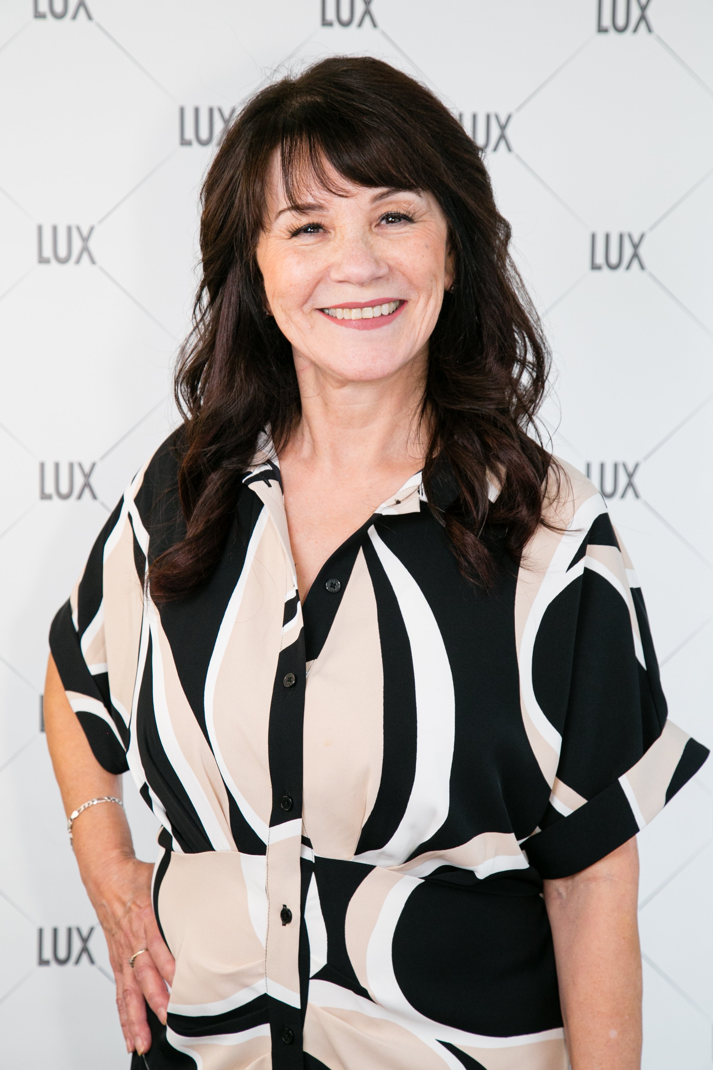 Smiling woman with dark brown hair wearing a black, beige, and white patterned blouse standing in front of a backdrop with the word "LUX" repeated.