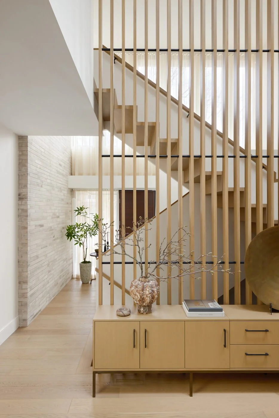 Interior view of a modern home with a wooden staircase and vertical wooden slats, a light-colored wood sideboard with decorative objects, and potted plants near a window with sheer curtains.