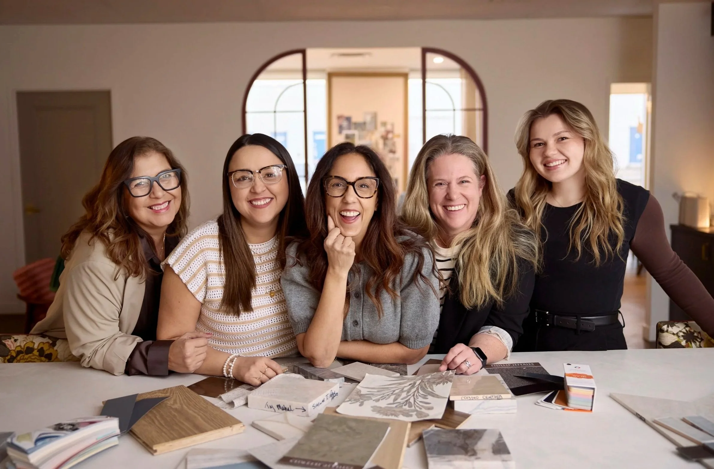 Nyla Free Designs team smiling and posing together behind a table with interior design samples and swatches in a well-lit room.
