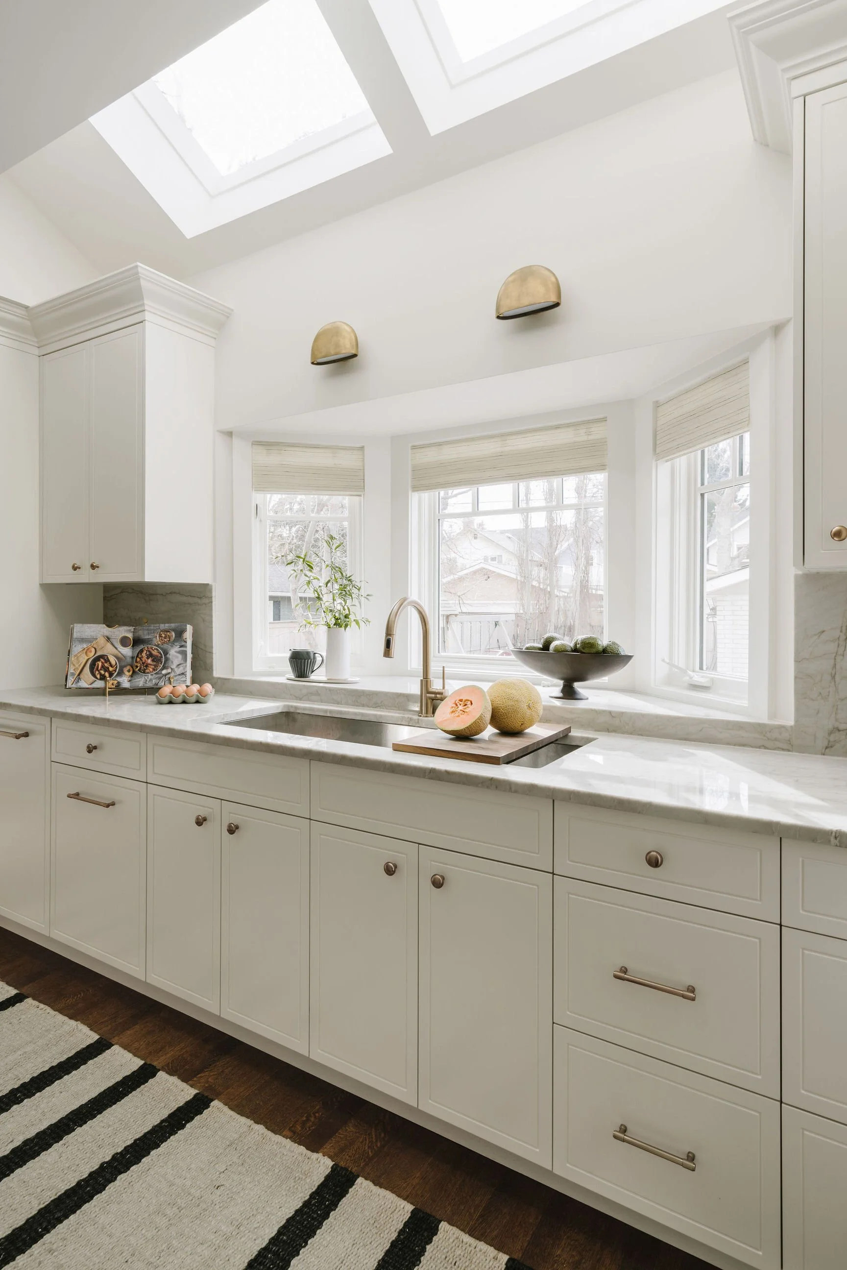 Bright kitchen with white cabinetry, marble countertops, and large windows. Sunlight streams through skylights. Decor includes a plant, a bowl of green fruit, and halved cantaloupe on a cutting board.