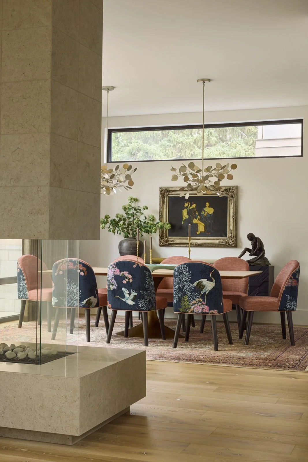 Modern dining room with a long table, eight upholstered chairs with floral and bird patterns, a decorative fireplace, a large painting, a sculpture, and greenery, illuminated by horizontal window and hanging light fixtures.