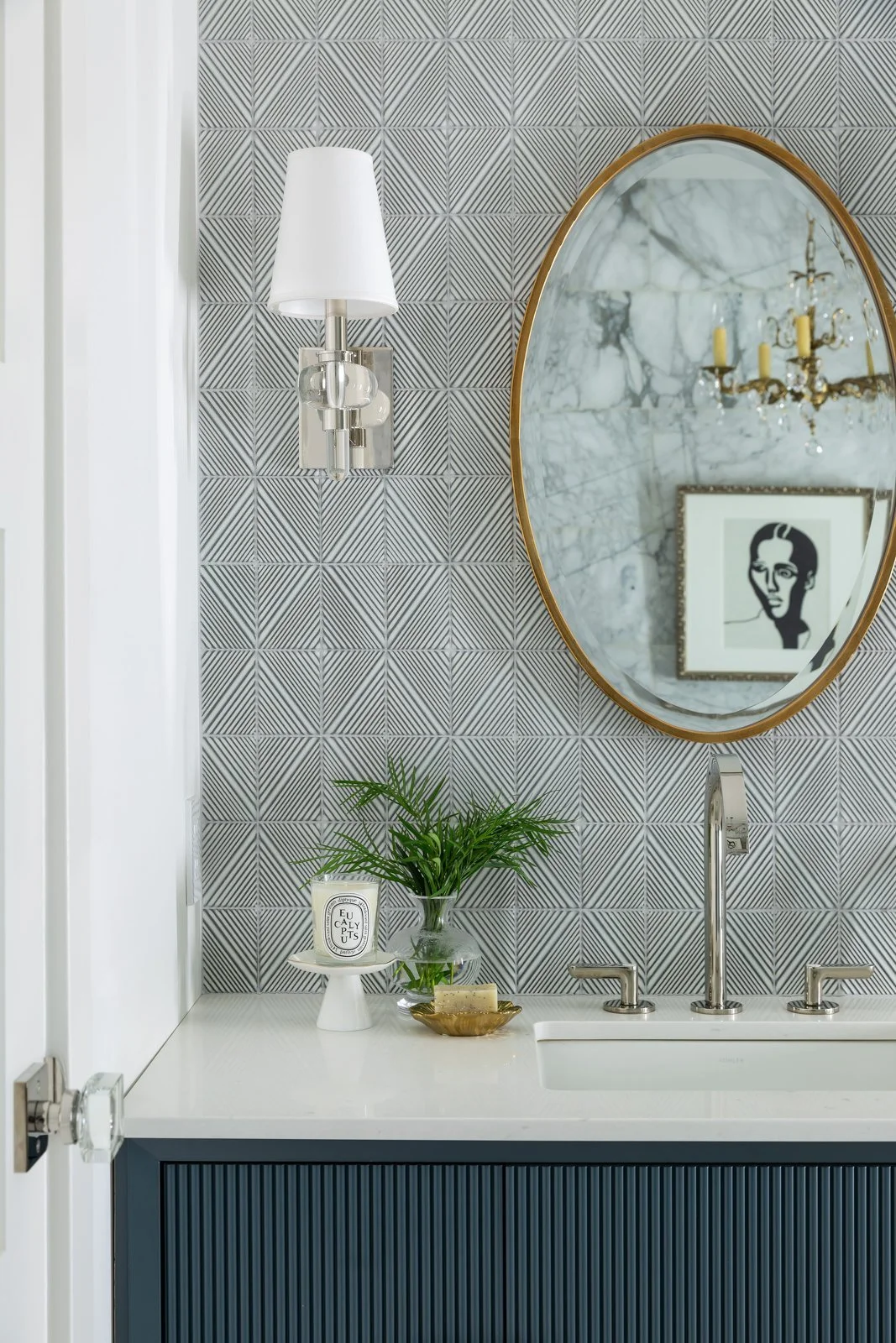 A bathroom vanity with a modern design featuring a white countertop, dark blue cabinet with vertical grooves, and a rectangular sink. Above, a round mirror with a gold frame reflects a chandelier and a framed art piece. To the left, a wall-mounted light fixture with a white shade and metallic base, and decorative items including a glass vase with green plant, candle, and soap dish.