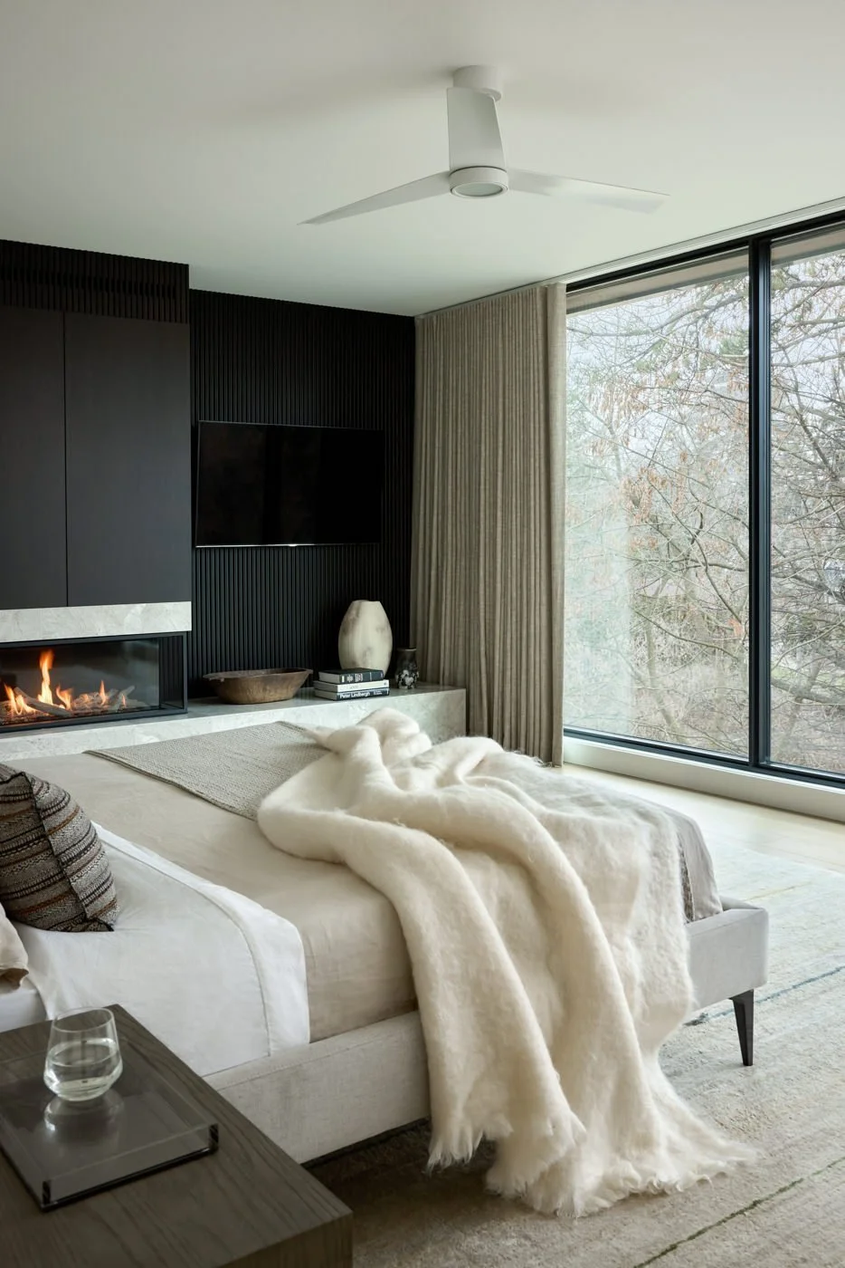 Modern living room with a cozy bed covered with a white faux fur blanket, a side table with a glass of water, a fireplace, a large window with beige curtains, a mounted TV, and decorative objects on a white mantel.