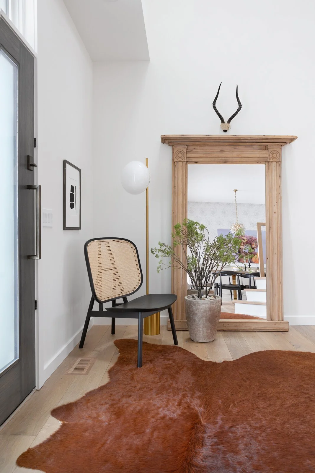 Interior entryway with a black wooden chair that has a woven cane backrest, a tall gold floor lamp with a white globe, a large potted plant, a wooden archway frame with a mirror, a brown cowhide rug, and a wall-mounted antelope skull with horns.