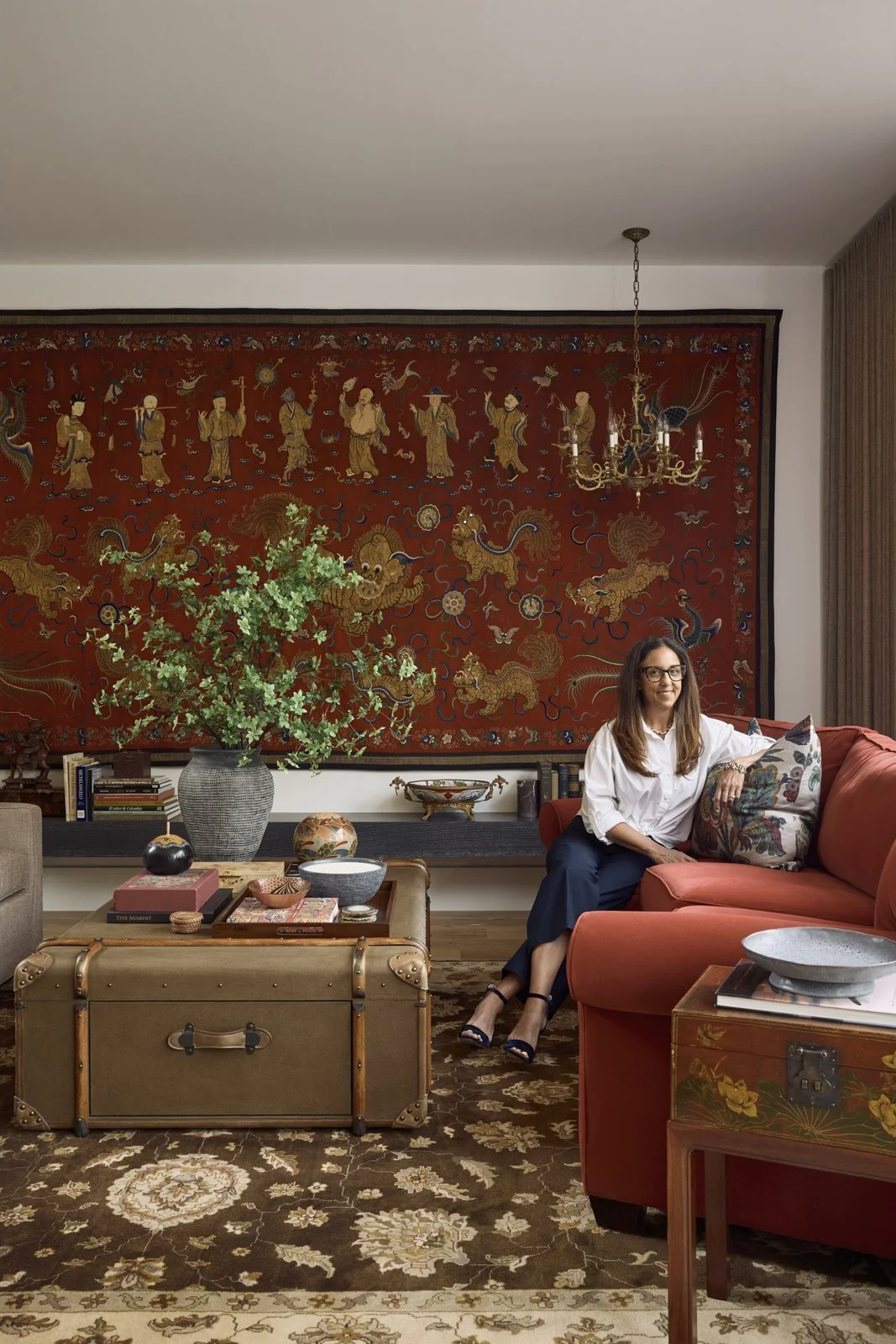 A living room with chic decor, including a large red tapestry with gold figures, a chandelier, a woman sitting on a red sofa, a coffee table with decorative items, and a large potted plant.