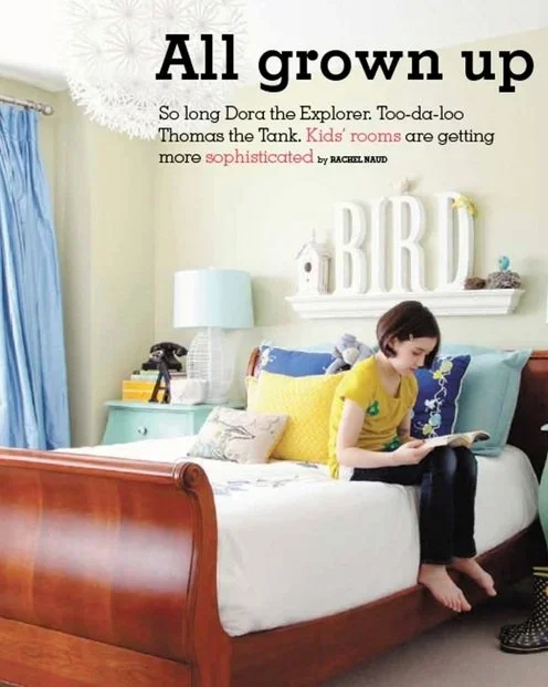 A young boy in a yellow T-shirt sitting on a bed, reading a book in a bedroom decorated with blue and yellow accents, including a nightstand with a lamp, a shelf with decorative letters spelling "BIRD," and a blue curtain.