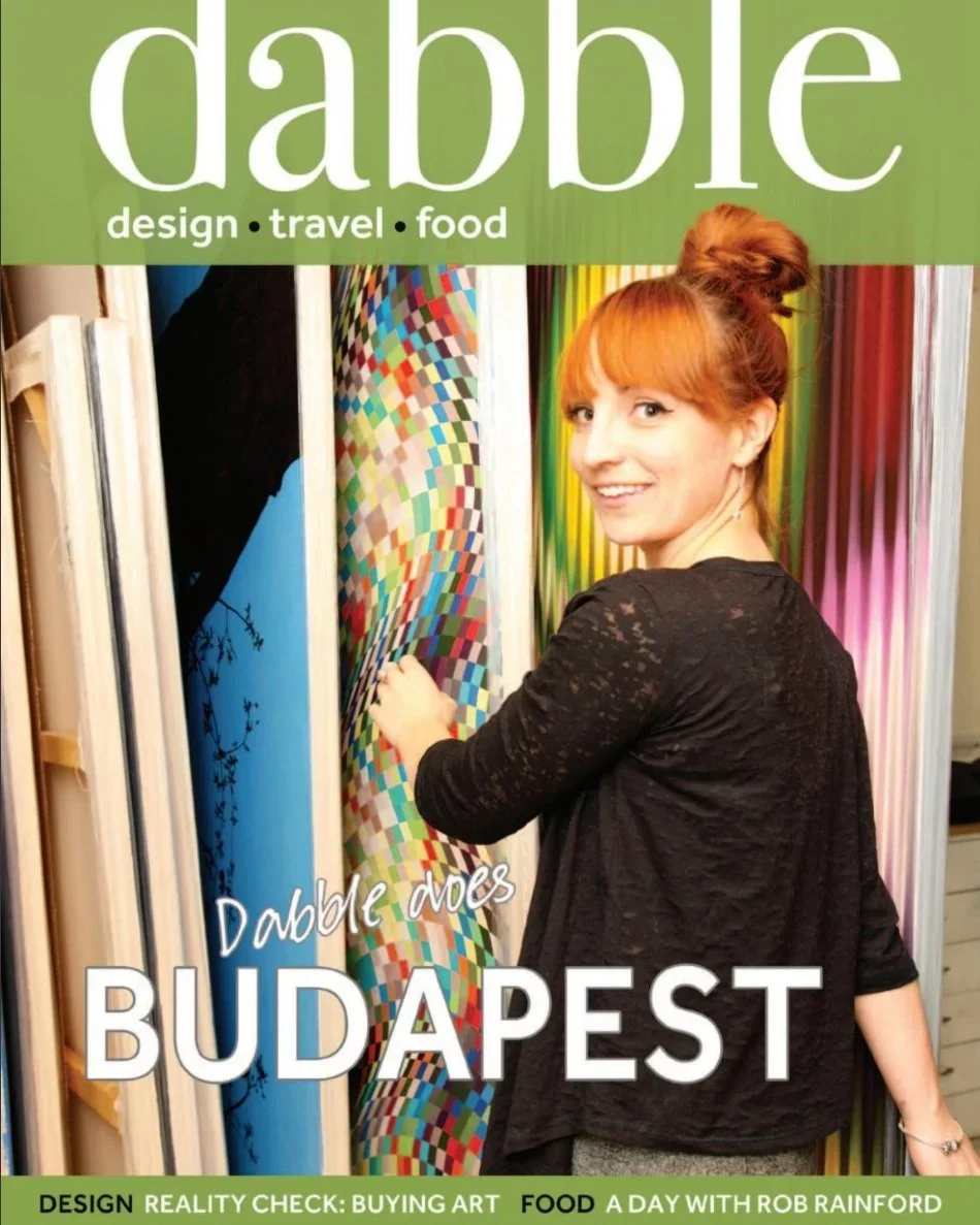 Woman with red hair in a bun smiling and touching colorful patterned panels at an art display. Cover of Dabble magazine with the title "Dabble does BUDAPEST" and topics "design, travel, food."