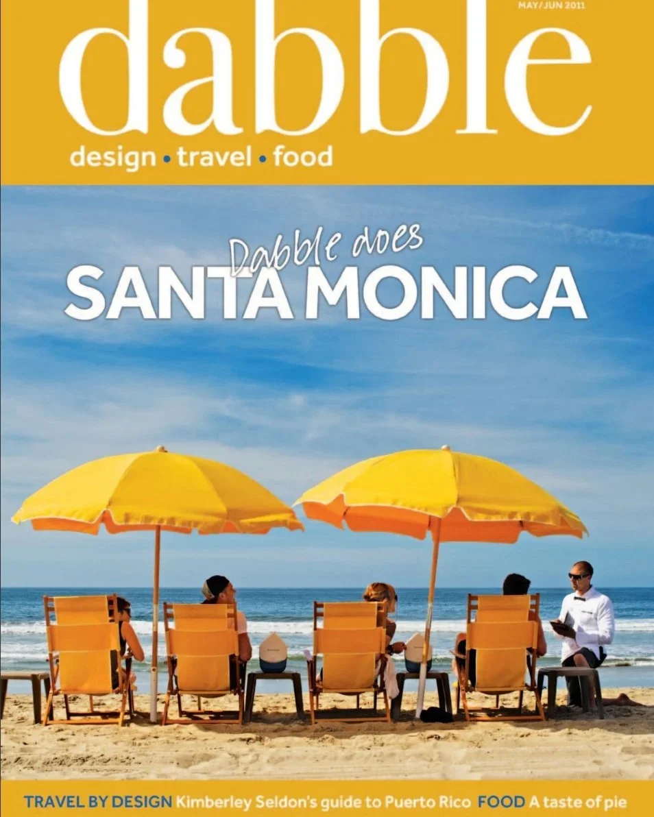 A magazine cover titled 'dabble' with a subtitle 'design, travel, food'. The cover features five people sitting on chairs on a sandy beach, facing the ocean with waves. Two yellow umbrellas shade the group. The magazine headline says 'Dabble does SANTA MONICA'.