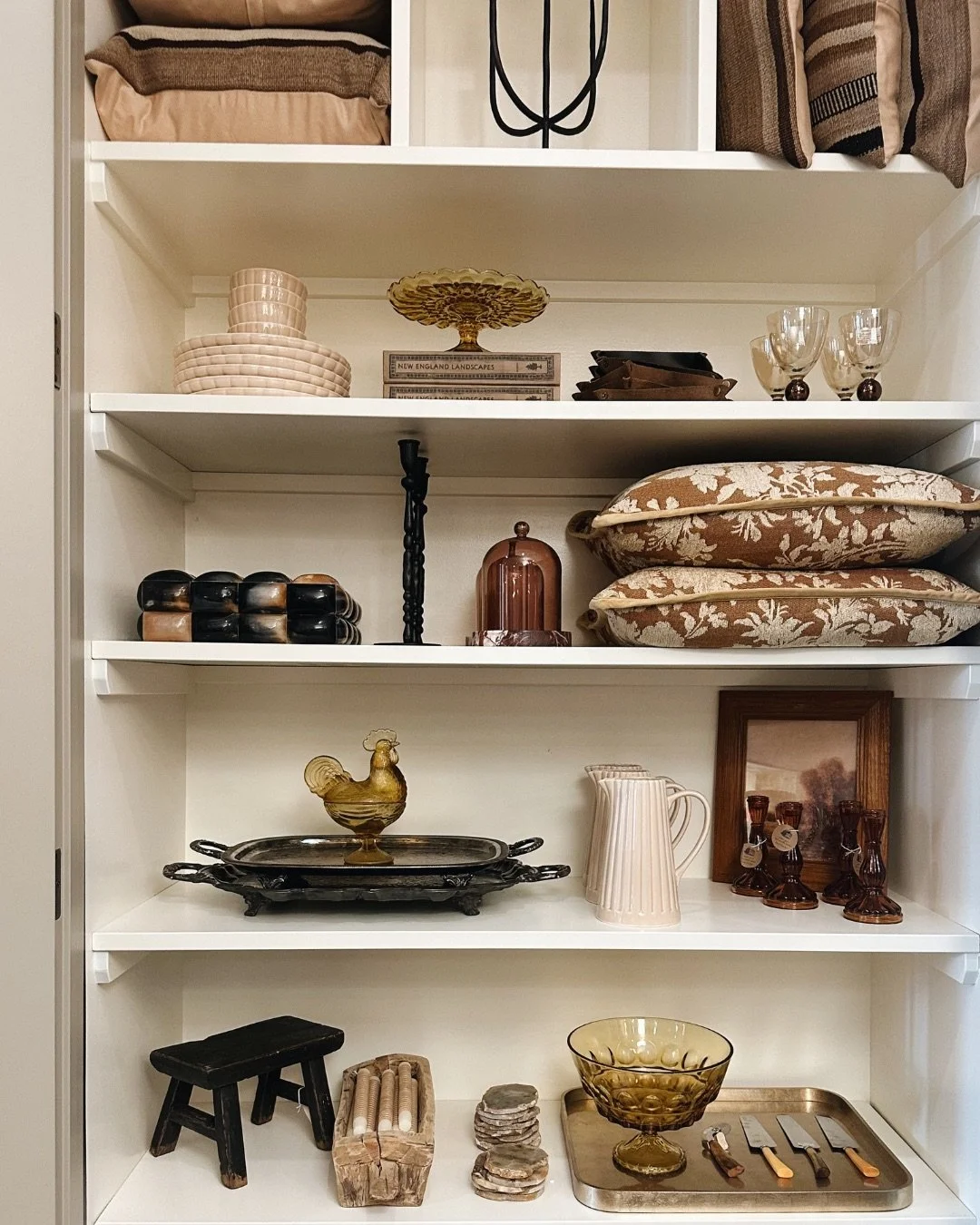 White shelving filled with decorative and vintage items including plates, bowls, glassware, small sculptures, framed pictures, and cutlery.