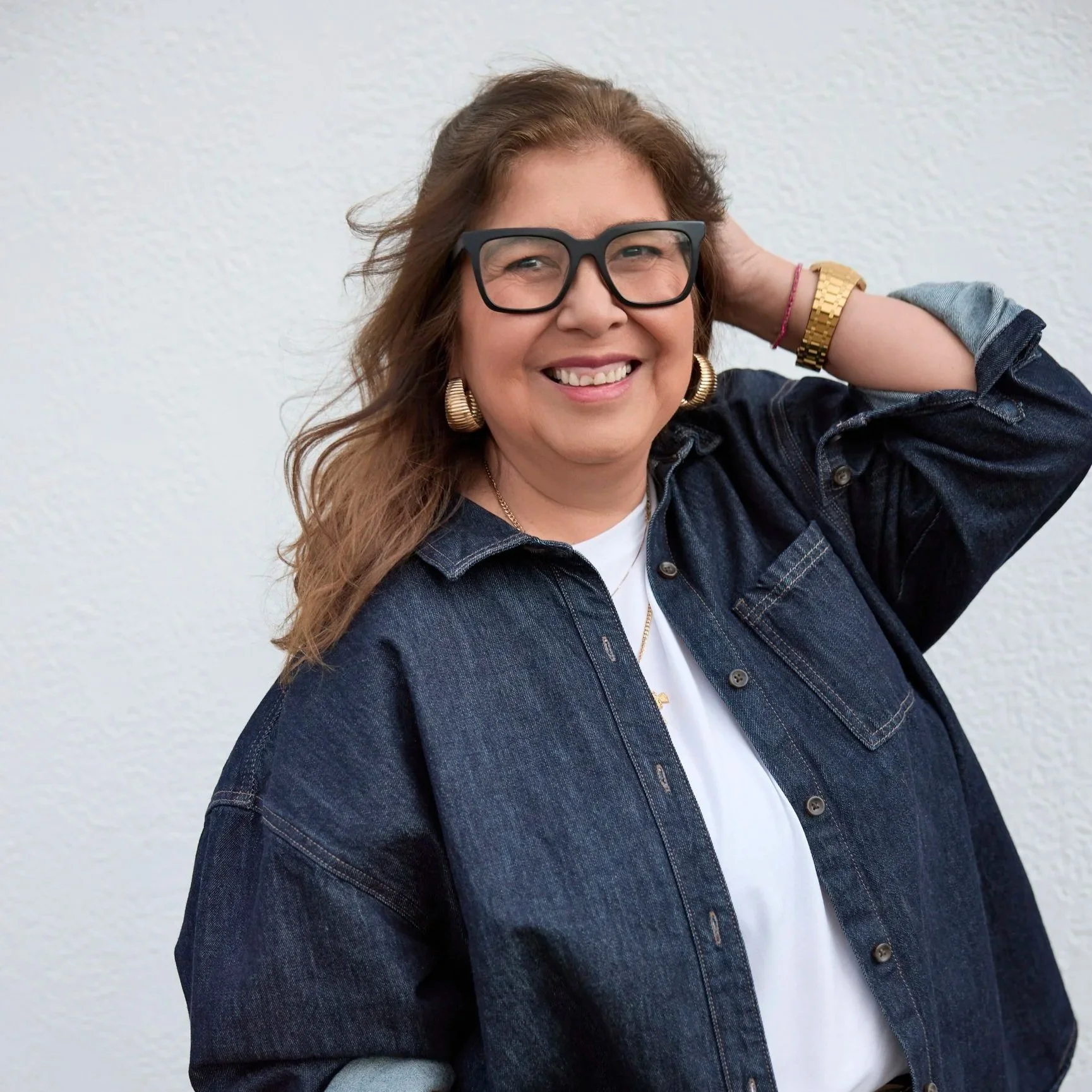 Woman smiling, wearing glasses, a denim shirt, and gold jewelry, standing against a white wall.