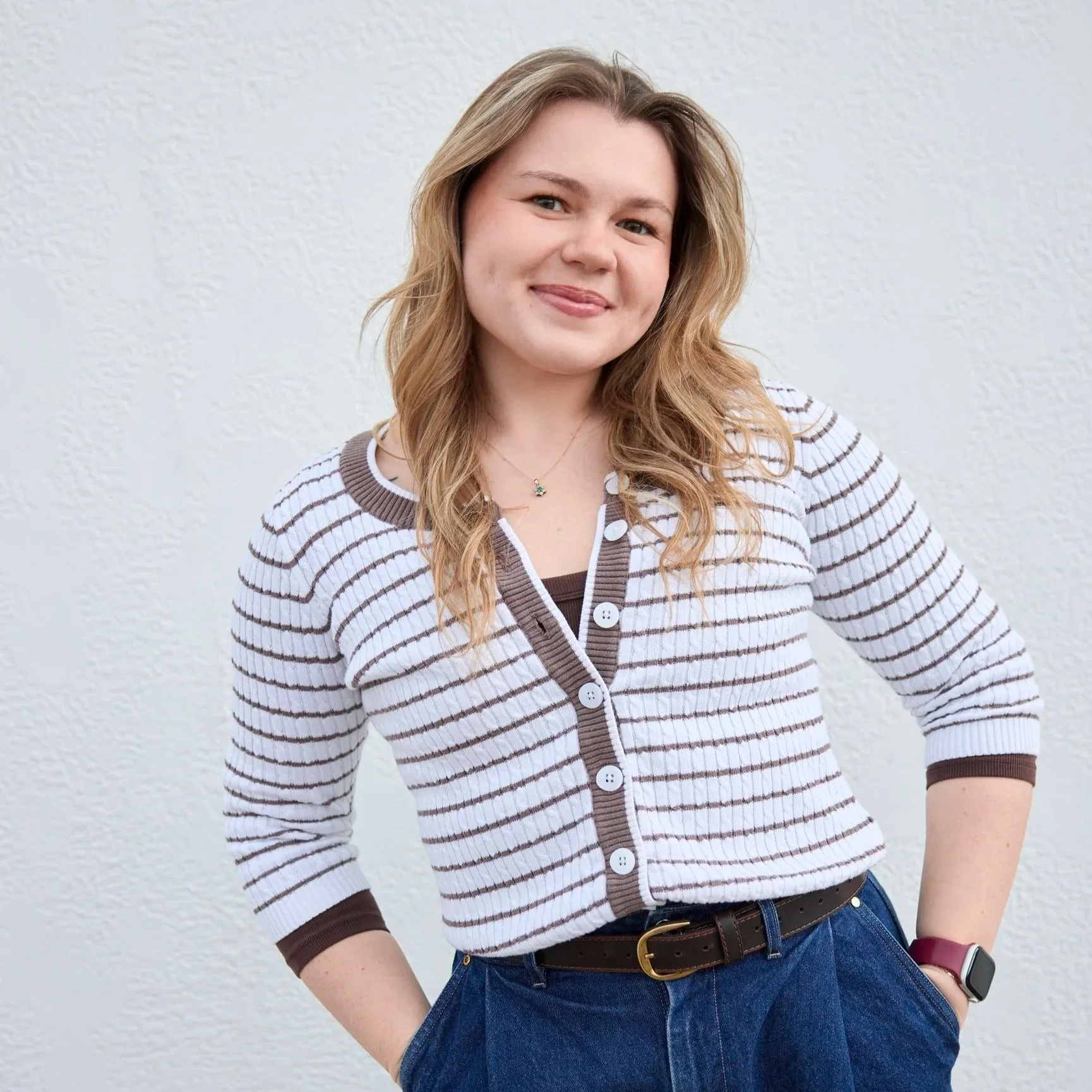 Young woman with light brown hair smiling, wearing a striped cardigan, dark top, jeans, and a smartwatch, standing against a light-colored wall.