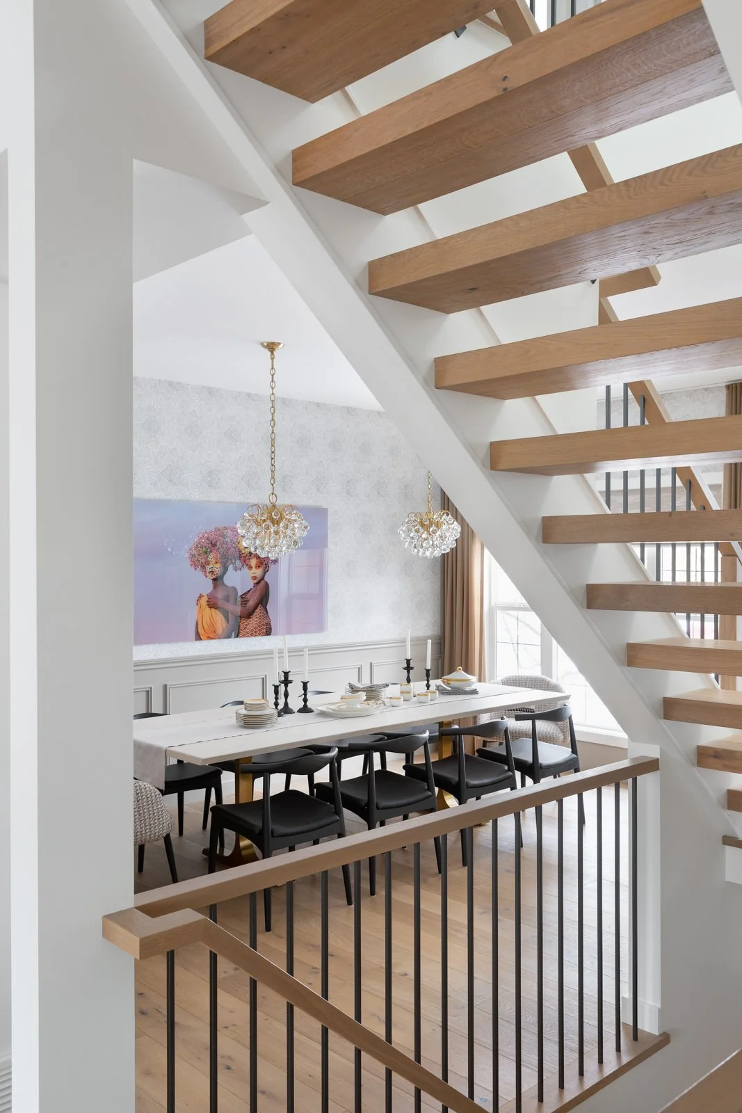 Dining room with a long white table set with plates, candlesticks, and cutlery, in a room with white walls, chandelier lighting, and black chairs, visible from the staircase area.