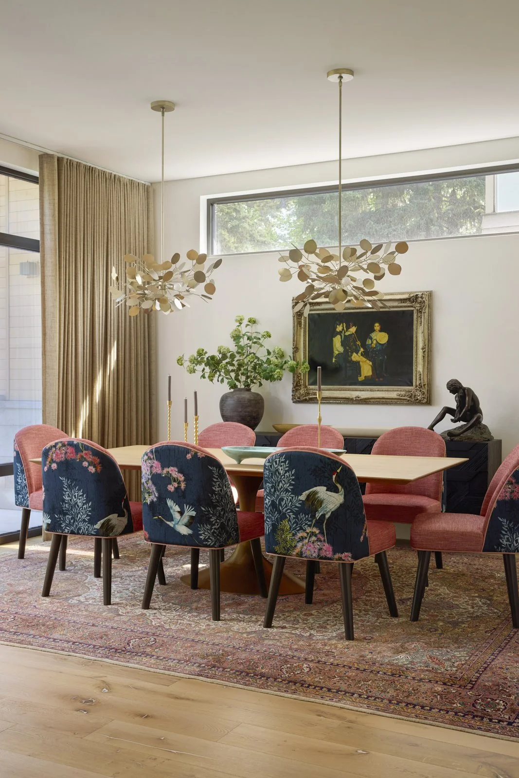 A modern dining room with a wooden table and ten chairs, some with floral and bird patterns, set on a vintage rug. The room features a large window, beige curtains, two contemporary chandeliers, a framed artwork, a large potted plant, and decorative statues.
