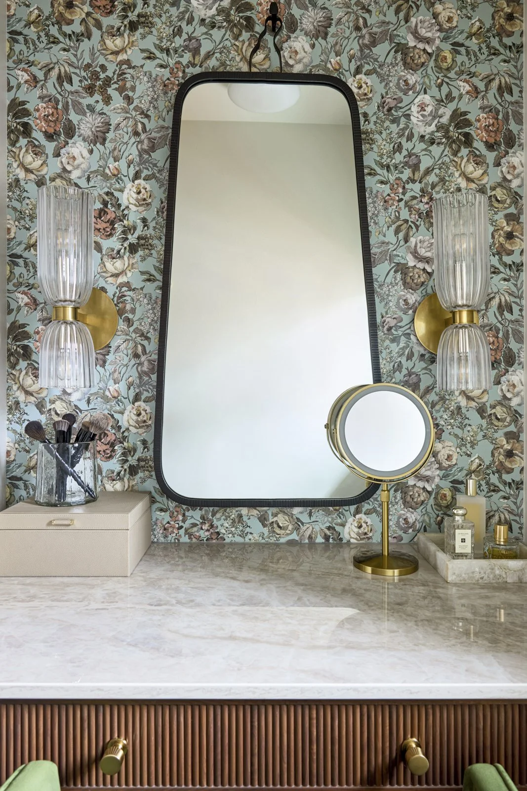 A bathroom vanity with a floral-patterned wallpaper, a mirror, two wall-mounted glass and brass lamps, a makeup mirror with a stand, and various bottles of perfume or toiletries on a marble surface.
