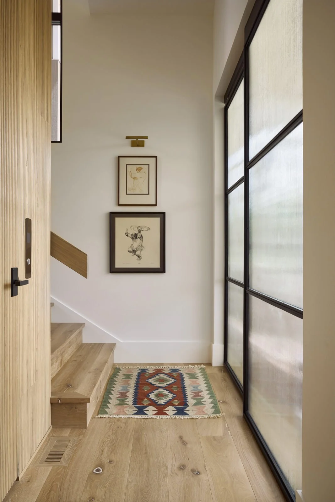 Entryway of a modern home with wooden stairs, framed artwork on white wall, a small colorful rug, and large frosted glass windows.