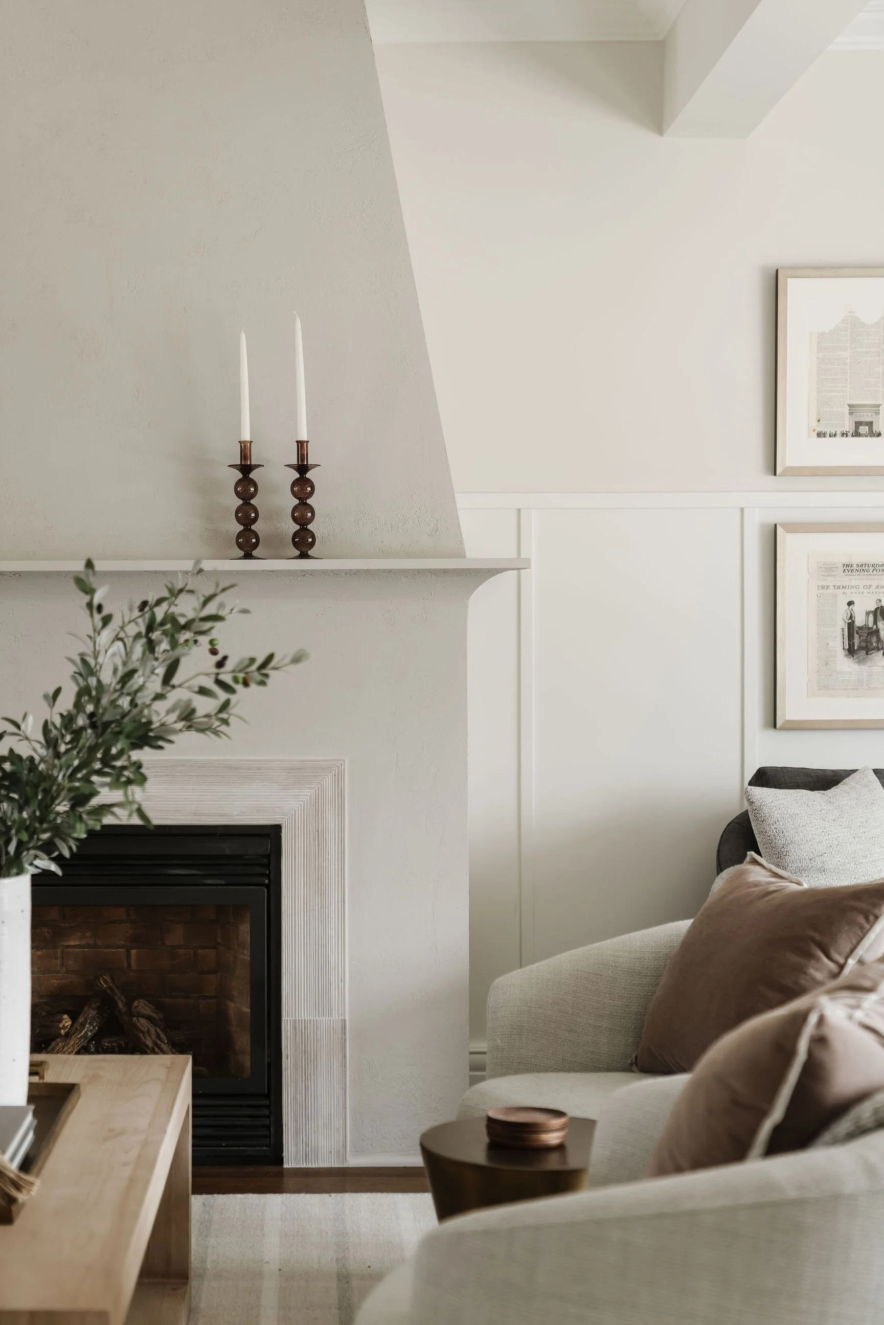 Living room with a fireplace, candles on the mantel, framed artwork on the wall, beige sofa with pillows, a wooden coffee table, and a vase with greenery.