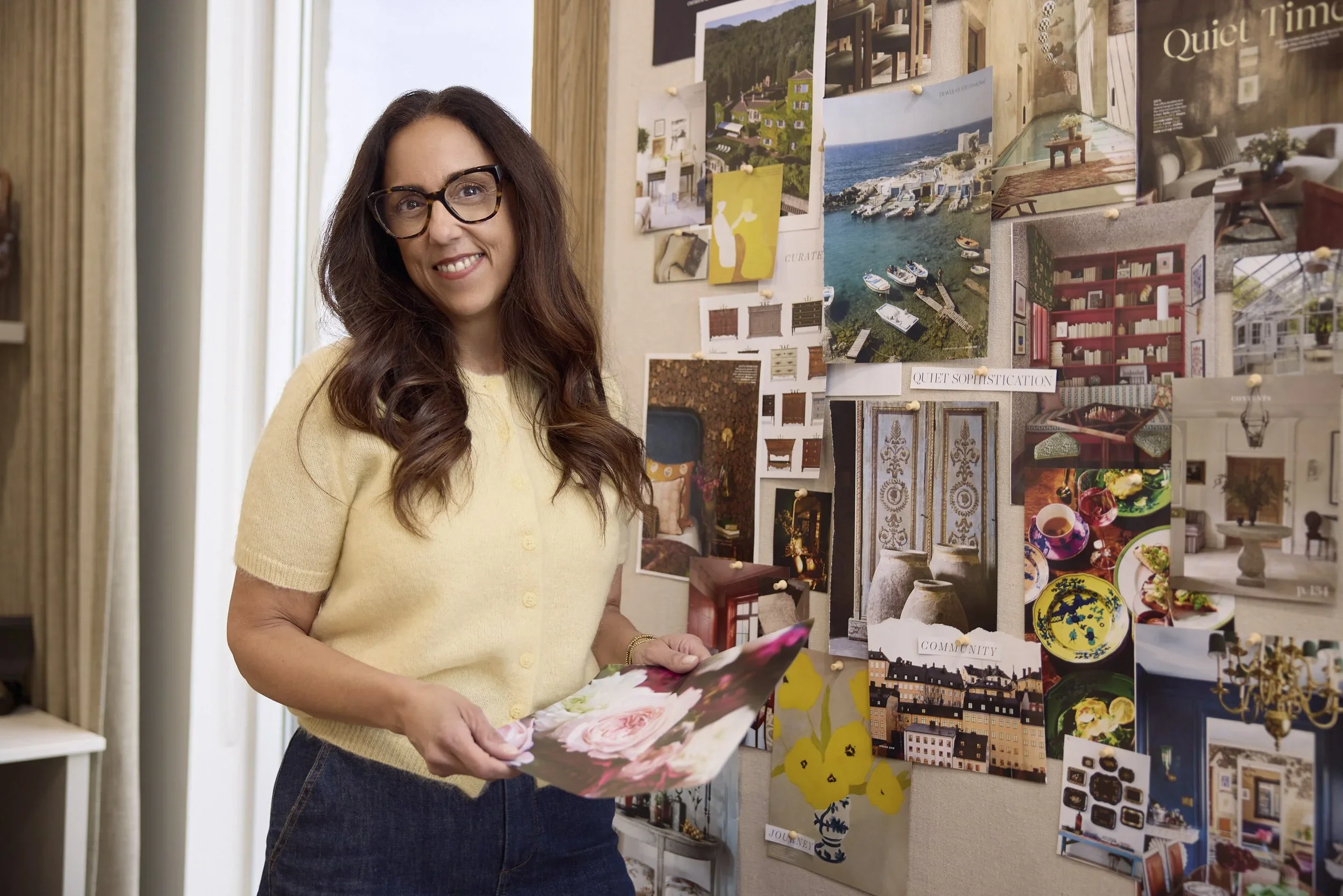 Woman with glasses and long brown hair smiling while looking at a pinboard filled with interior design and scenery photos, holding a floral photo album.