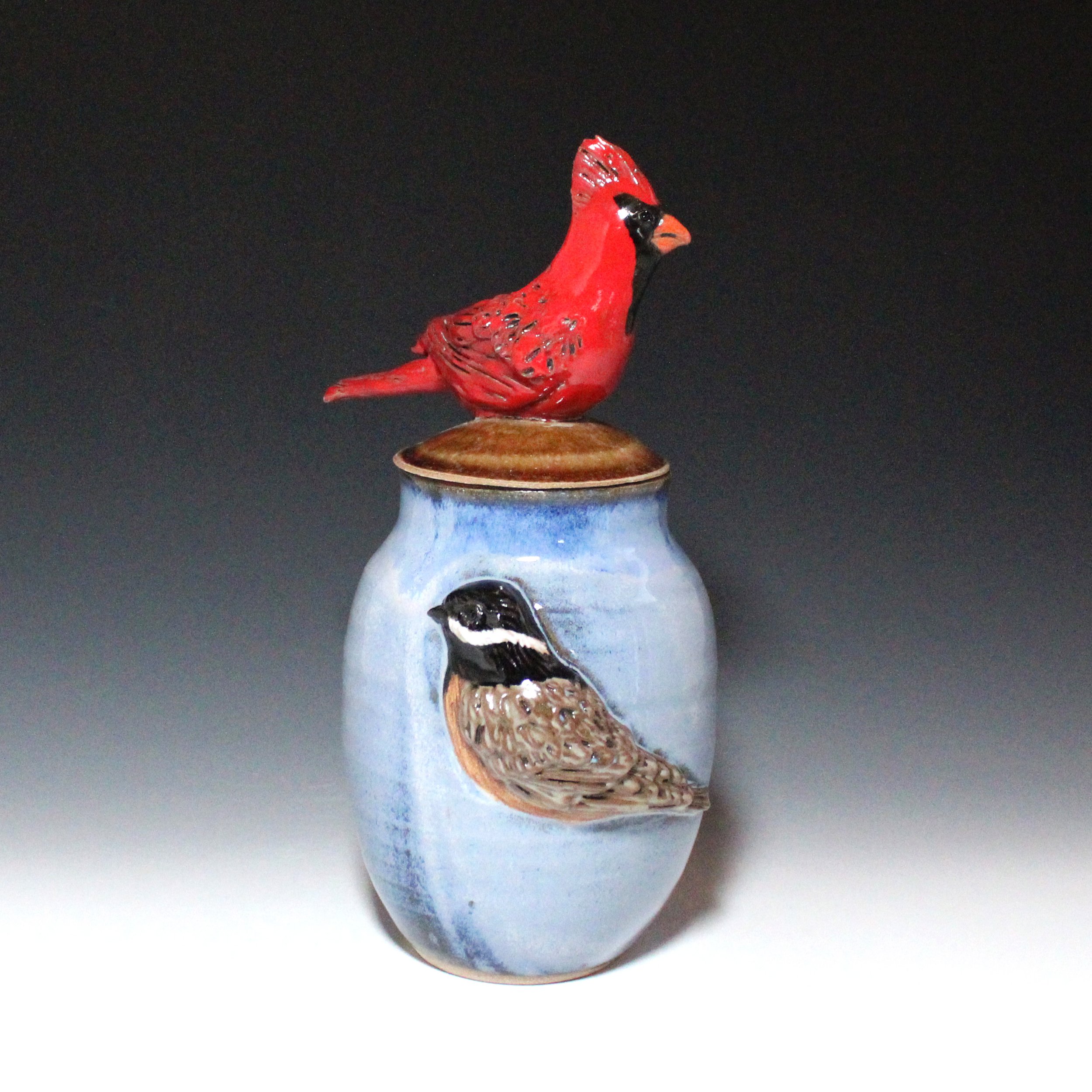 Original cardinal sculpture art figurine is perched on on the lid of the blue and brown lidded pot or urn. The side of the pot features an original chickadee relief.