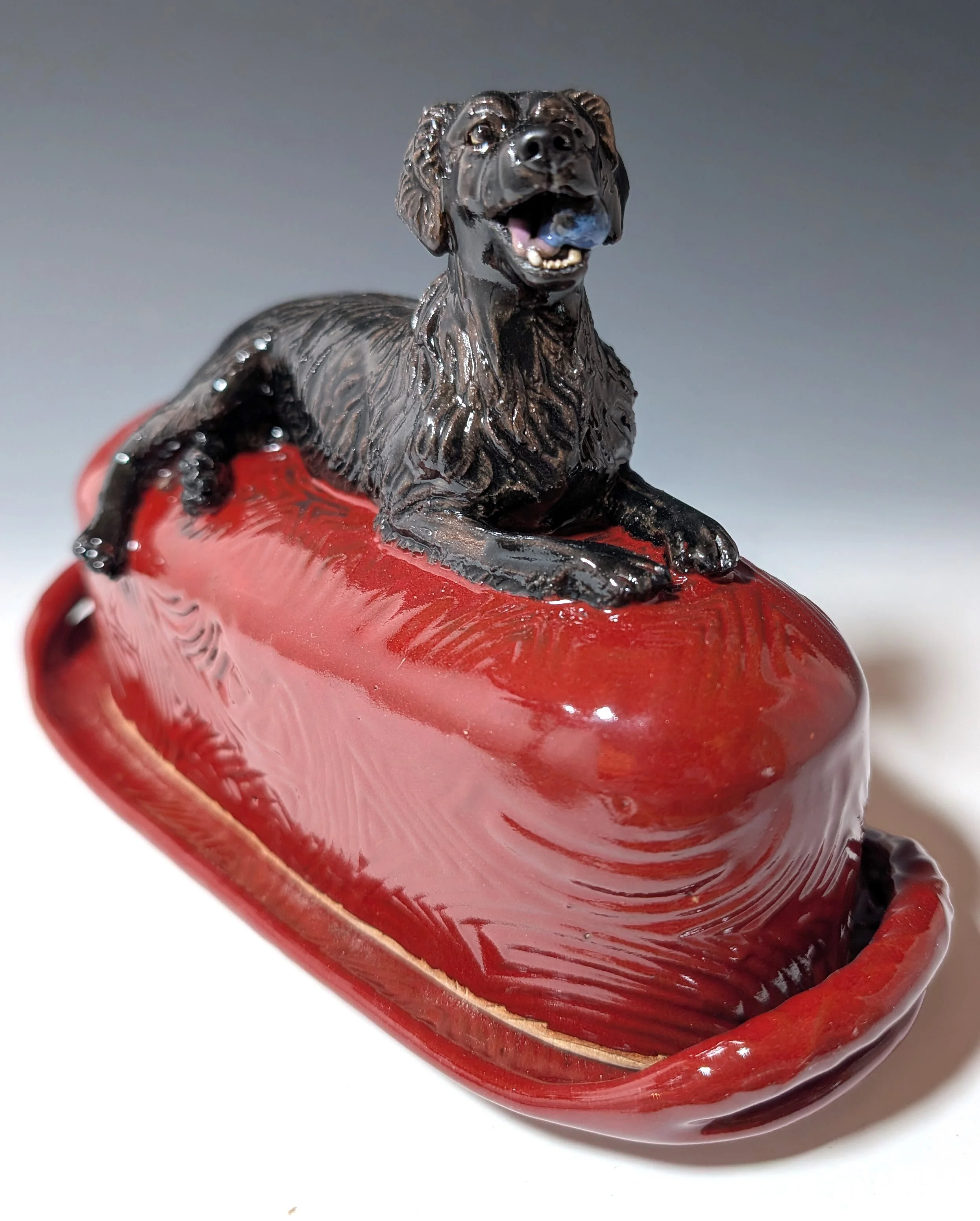 A handmade ceramic butter dish and lid glazed in red and featuring a custom sculpture of a black Labrador laying across the top and holding a blue ball in its mouth