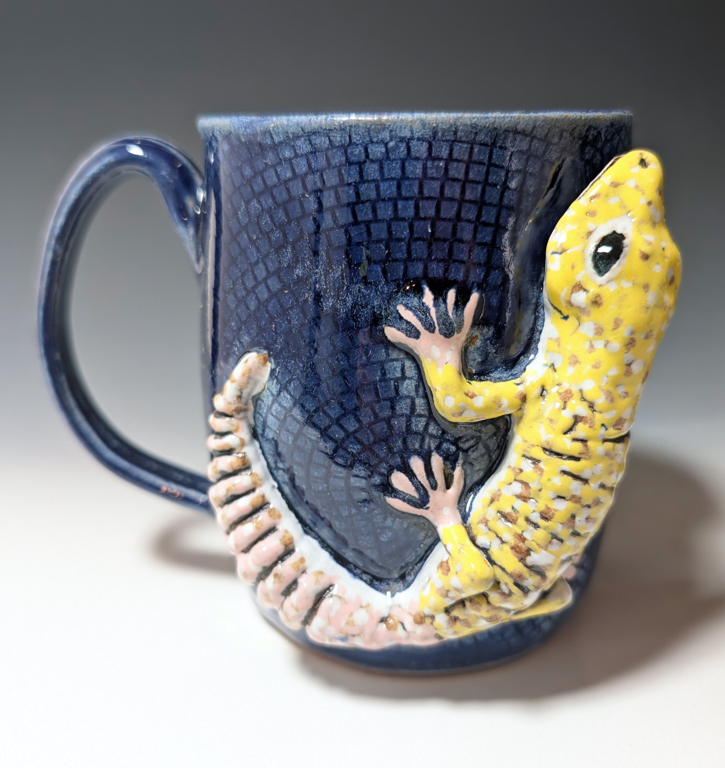 Blue handmade ceramic mug with a geometric texture and a large yellow and pink leopard gecko sculpture on one side.
