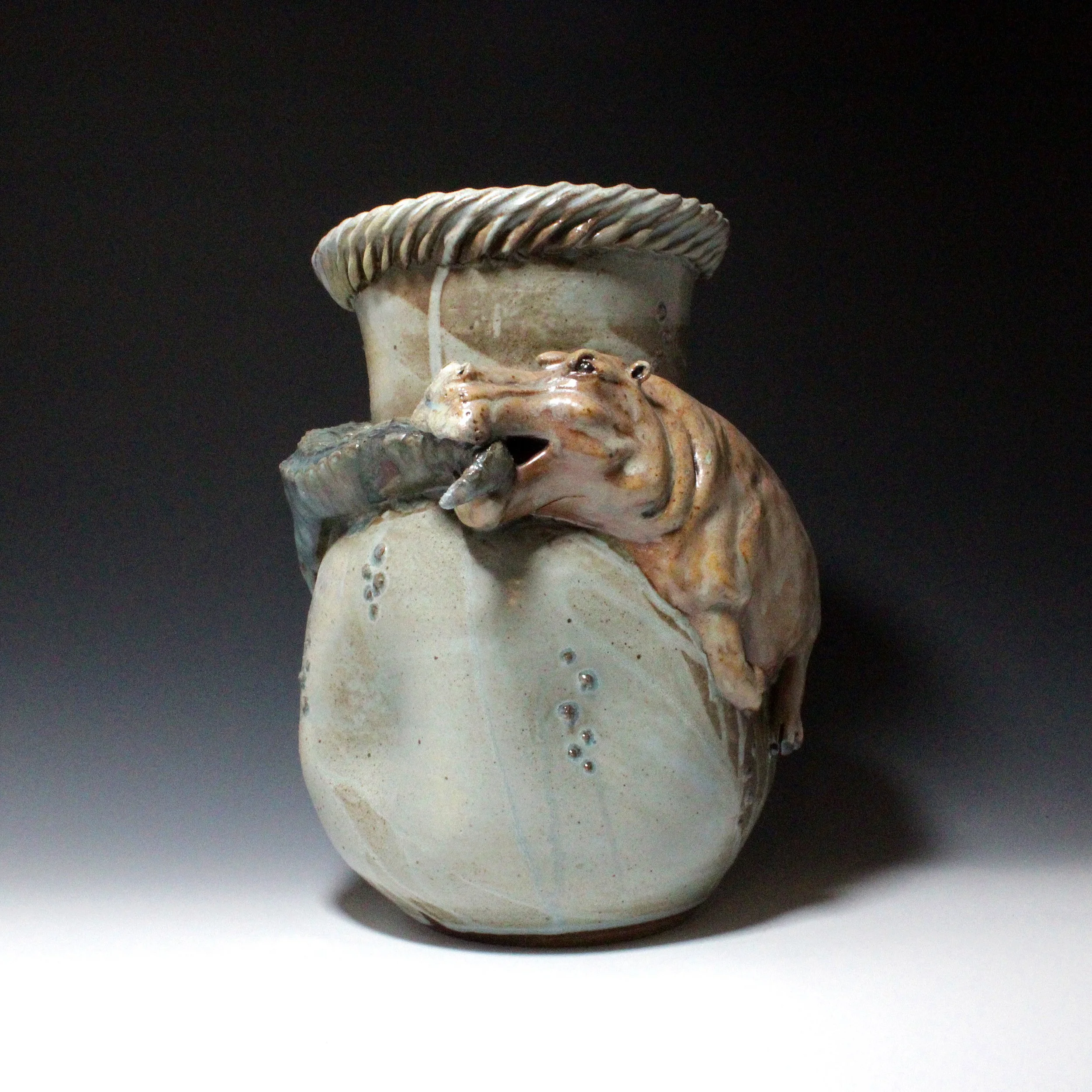 A large brown hippopotamus does not intend to share its watery home on the side of this blue vase. The hippo defends territory against an invading glassy green alligator by biting its tail.