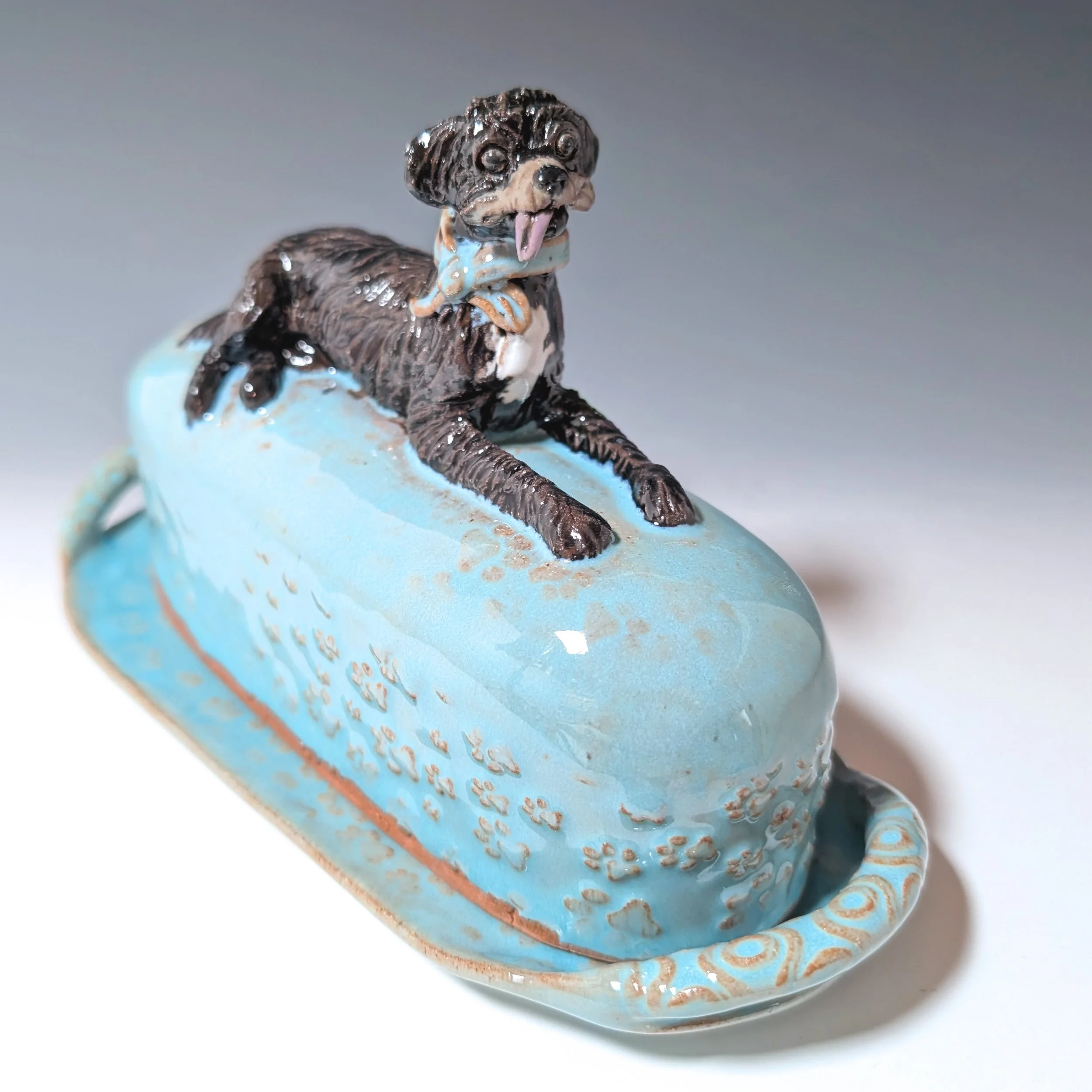 Bright blue lidded butter dish made from stoneware clay and featuring a hand sculpted dog sculpture on the lid. Dog figurine is laying across the top of the lidded butter plate with a blue bandana around its neck that matches the lidded butter keeper