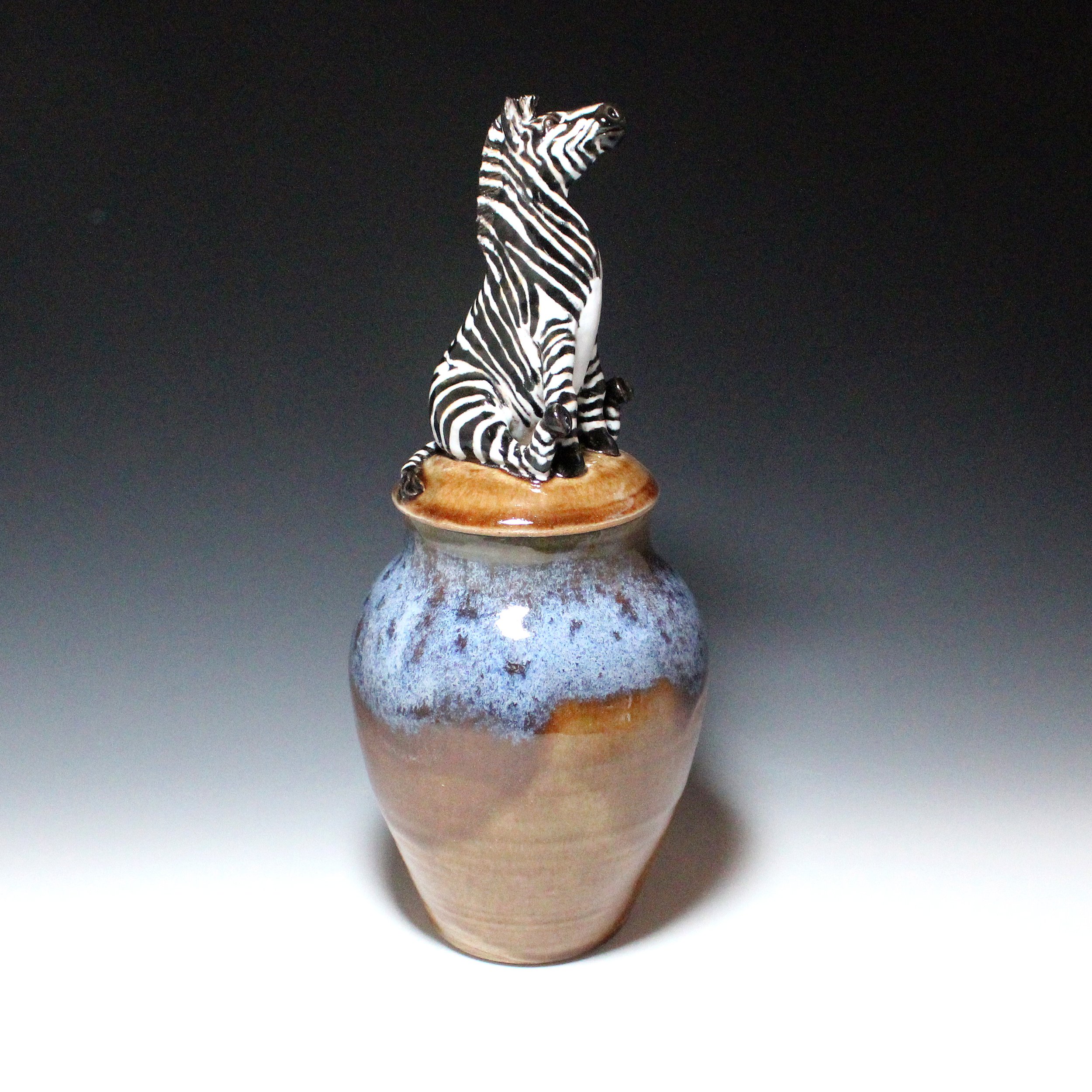 Hand sculpted zebra figurine sits on its rump on the lid of this brown and blue lidded pot, or urn.