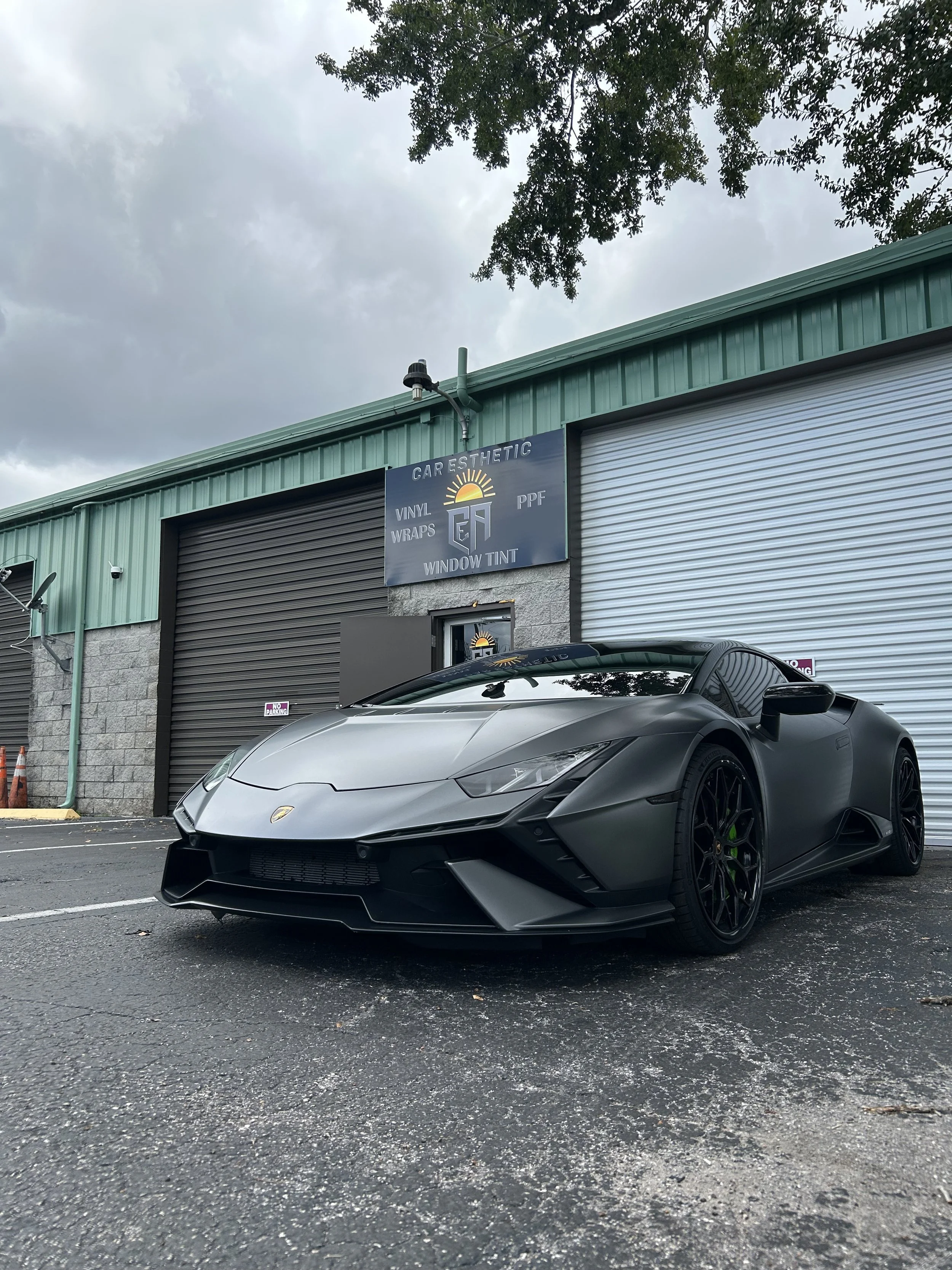 A matte black Lamborghini sports car parked in front of a commercial building with closed garage doors, a sign that reads 'Car Aesthetic' and services such as vinyl wraps and window tint, under a cloudy sky.