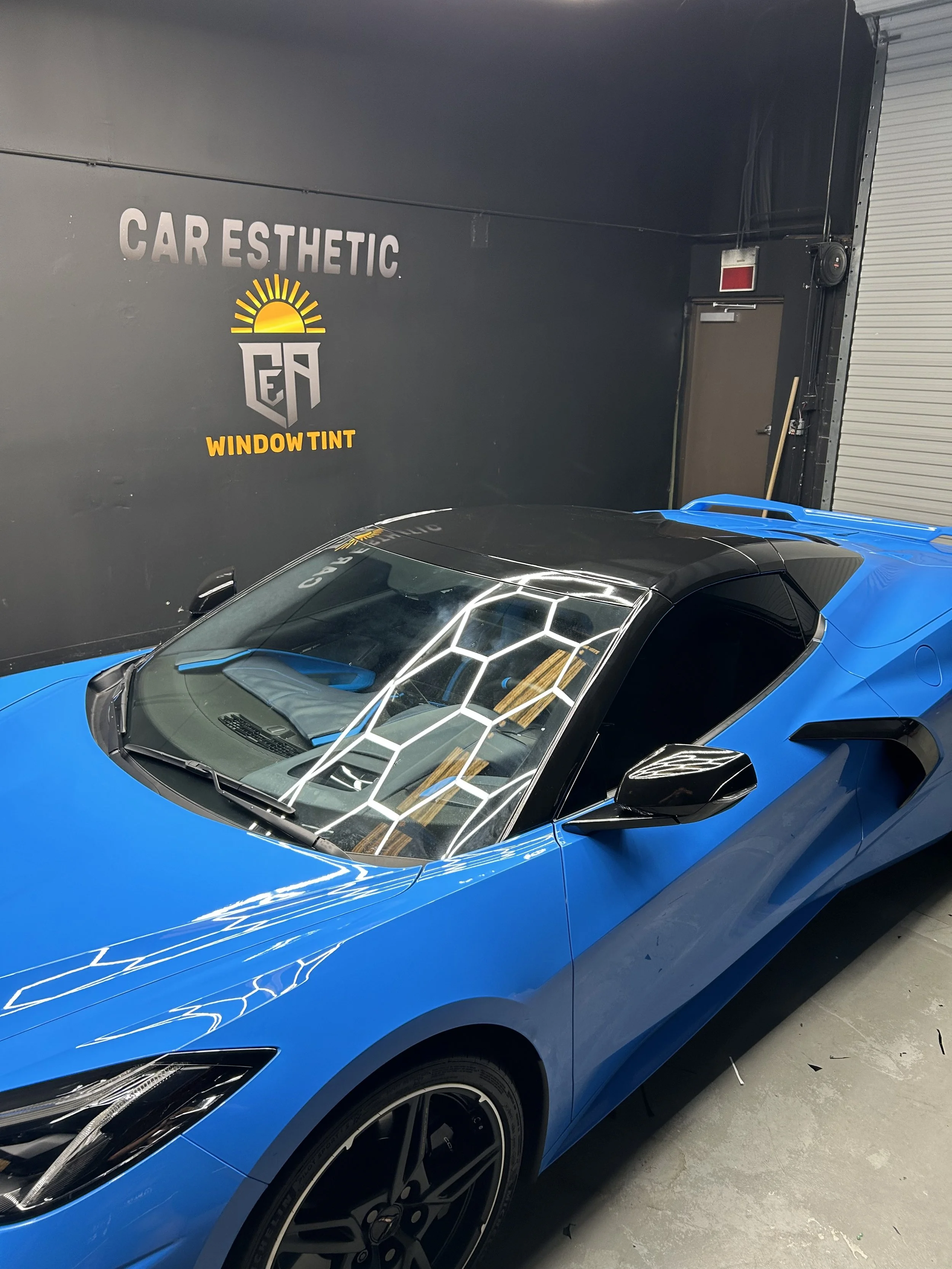 Blue sports car inside an automotive detailing shop with black walls labeled 'CARMESTHETIC WINDOW TINT'.