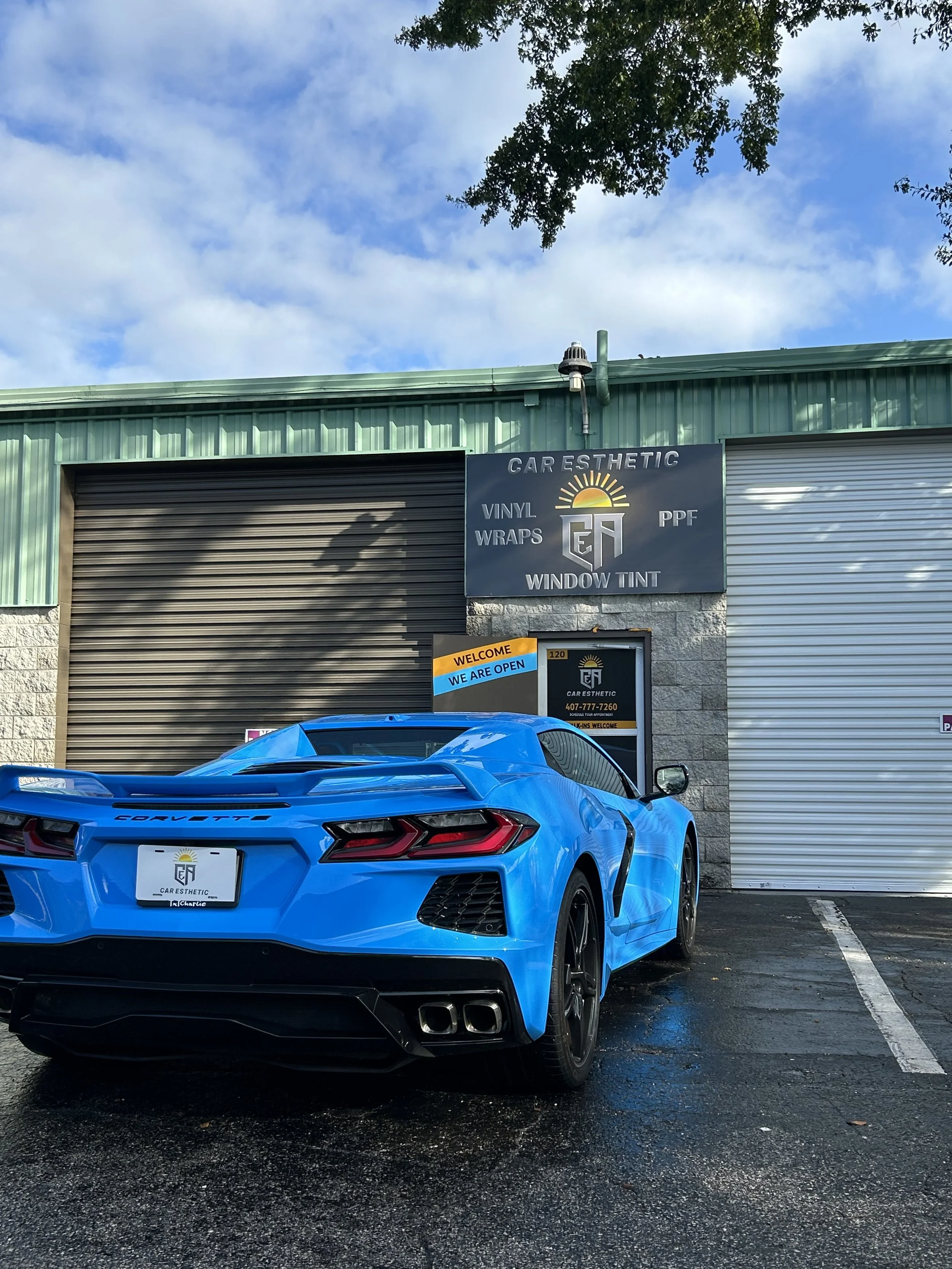 Blue Chevrolet Corvette sports car parked outside a car aesthetic shop with closed roller doors, a sign reading 'We Are Open,' and a building with green metal siding.