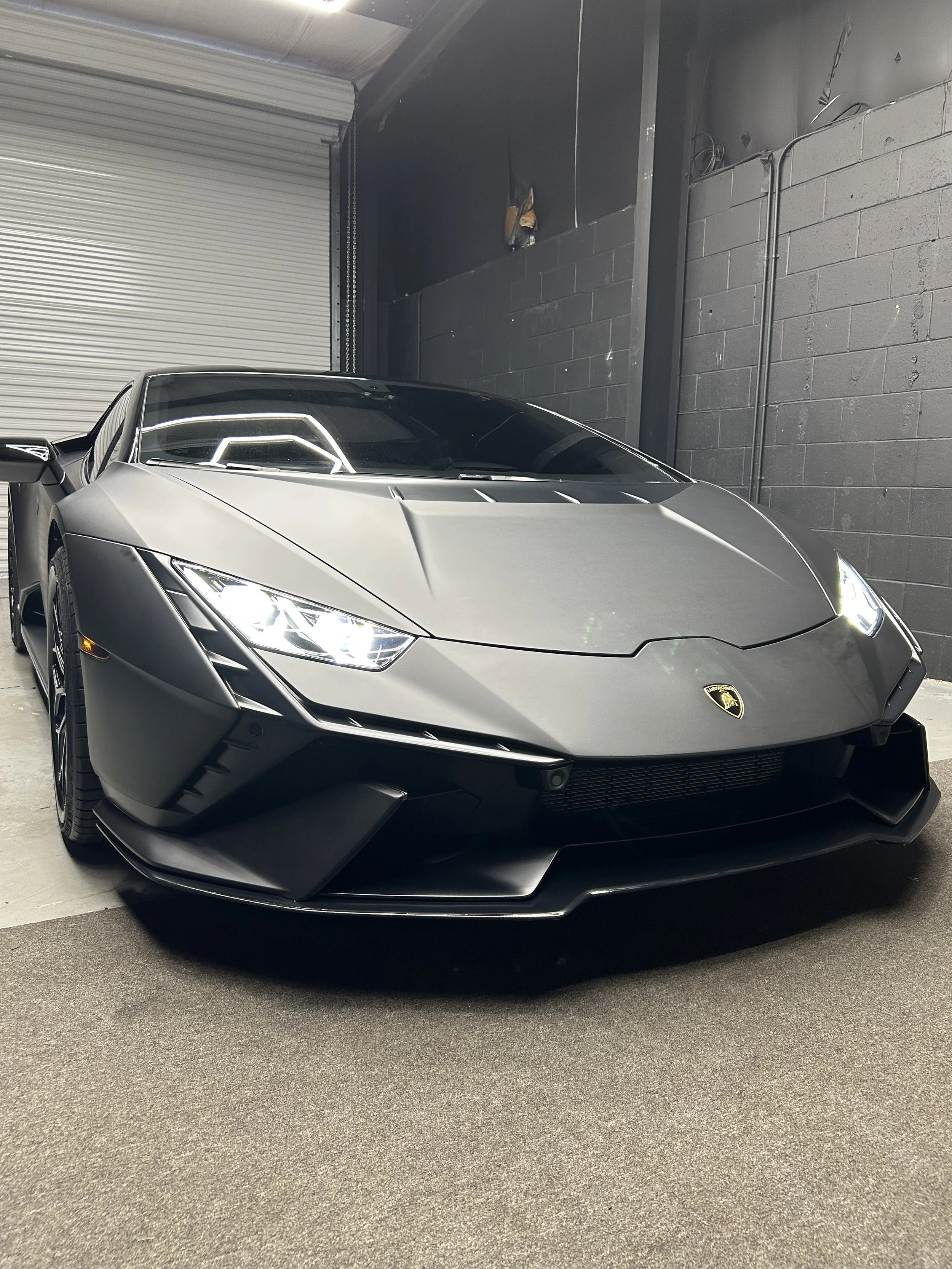 A silver Lamborghini sports car parked inside a garage with a gray concrete floor, black painted wall, and a partially visible metal roll-up door.